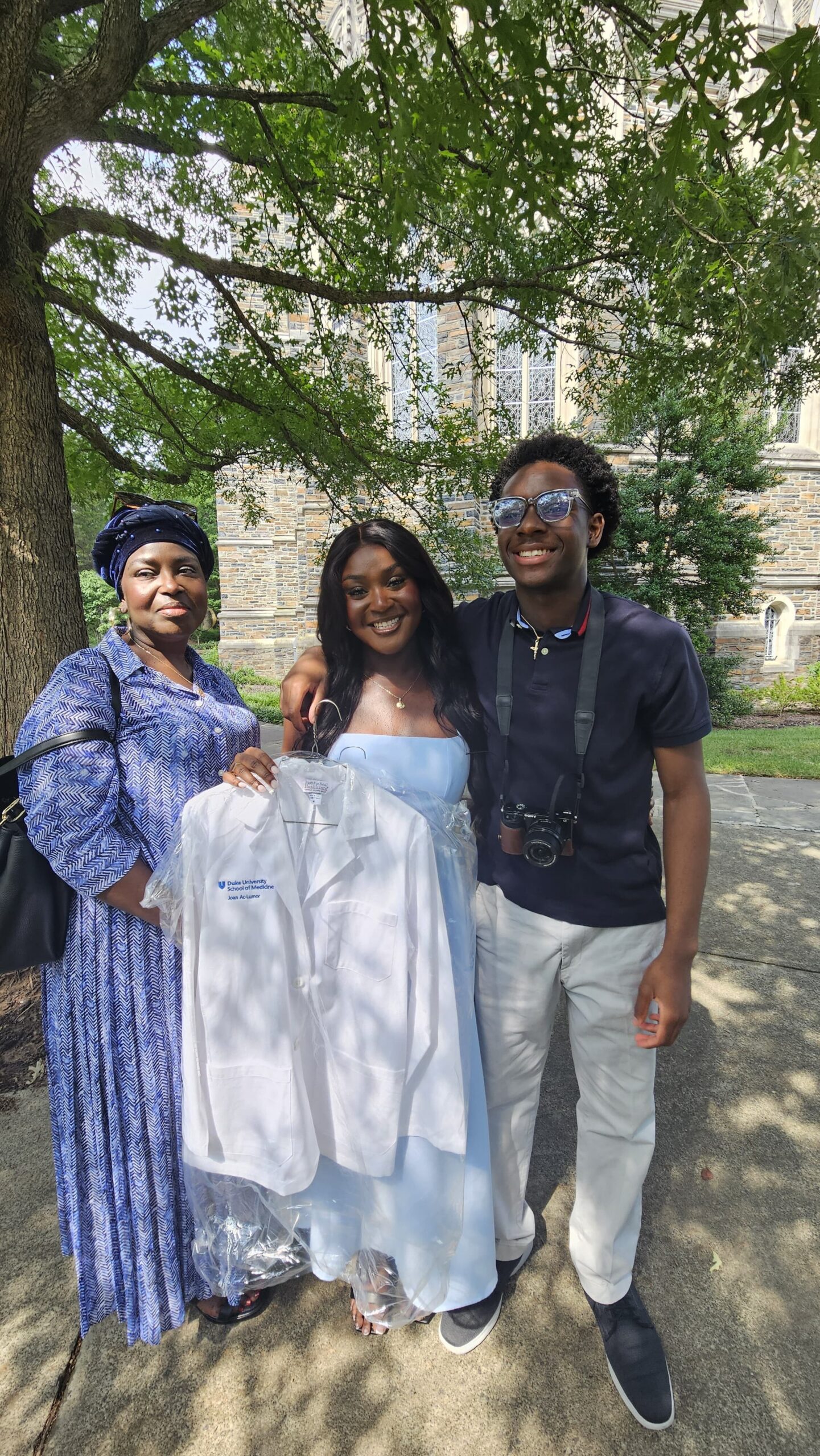 Three friends pose outside a stone building, with a woman in a blue dress and headwrap on the left, a woman in a light blue dress holding a white gown in the middle, and a man with a camera on the right.