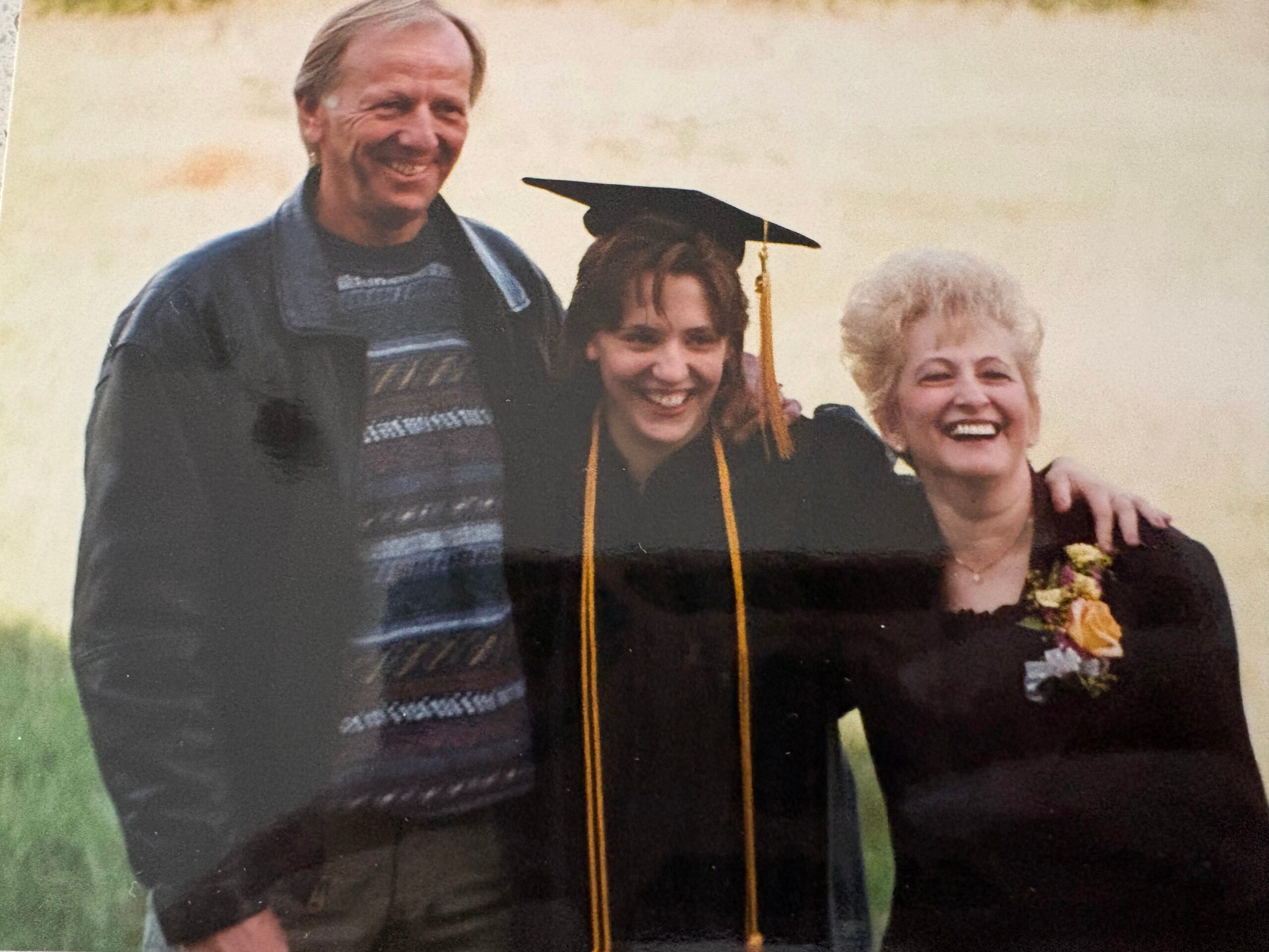 Three people pose outdoors for a graduation photo: the graduate in cap and gown with tassel, flanked by a smiling man and woman.
