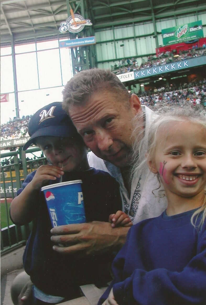 Man with two kids posing at a baseball stadium; boy holds a Pepsi cup while the girl smiles with face paint in a blue shirt.
