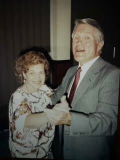 Smiling elderly couple dancing indoors; woman in a floral blouse, man in a gray suit with a red tie.