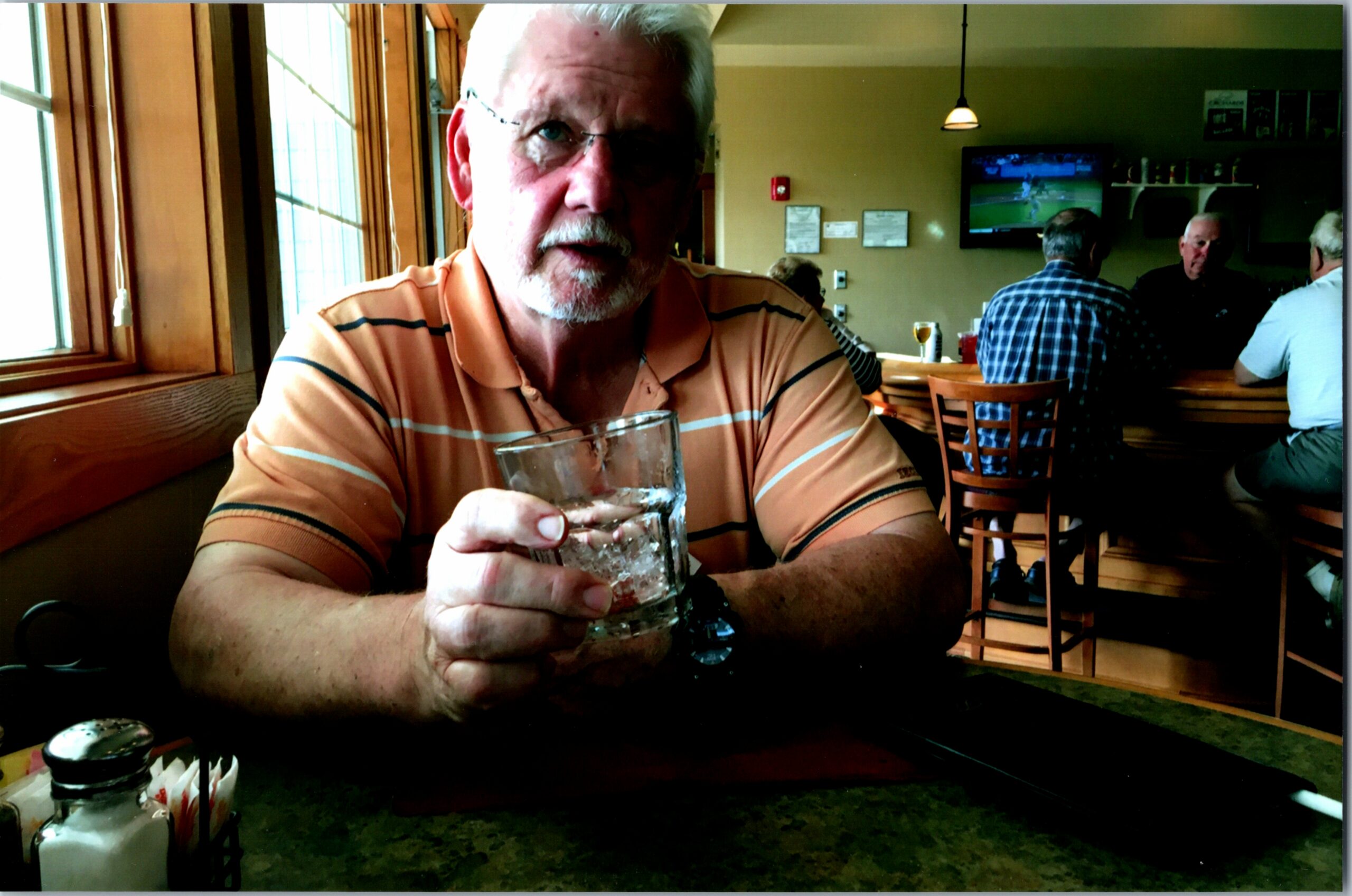Man with white beard in an orange striped polo holding a glass at a restaurant table, with a bar and TV in the background.
