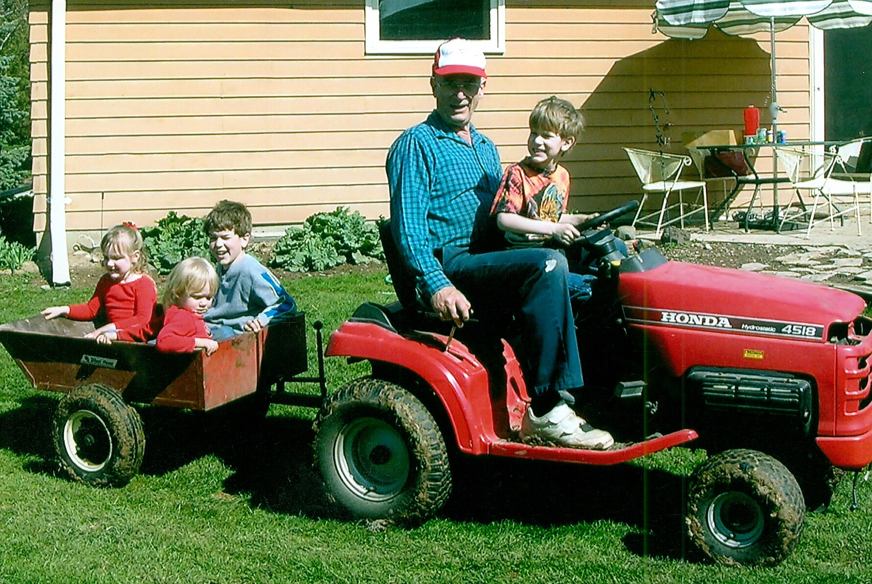 Man driving a red Honda garden tractor with three kids riding in a red wagon attached to the back, outdoors in a yard.