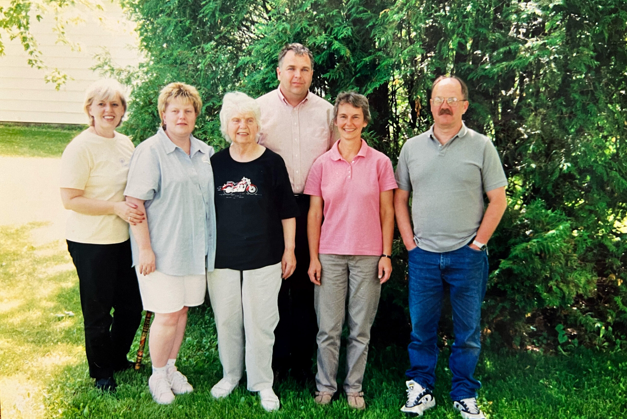 Six adults standing side by side outdoors, smiling, with trees and a lawn in the background.