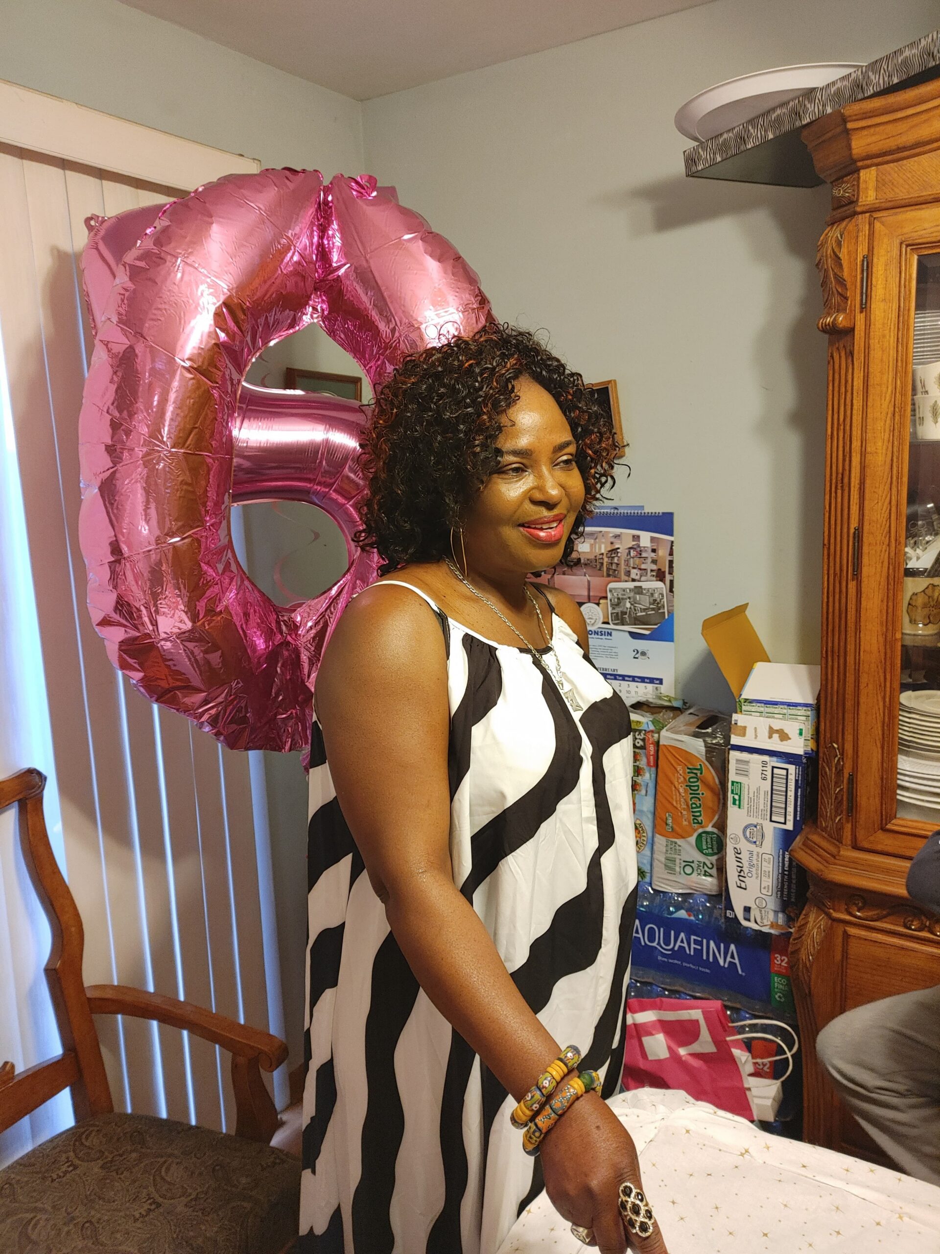 Woman in a black-and-white striped dress stands indoors with a large pink metallic birthday balloon behind her, smiling.