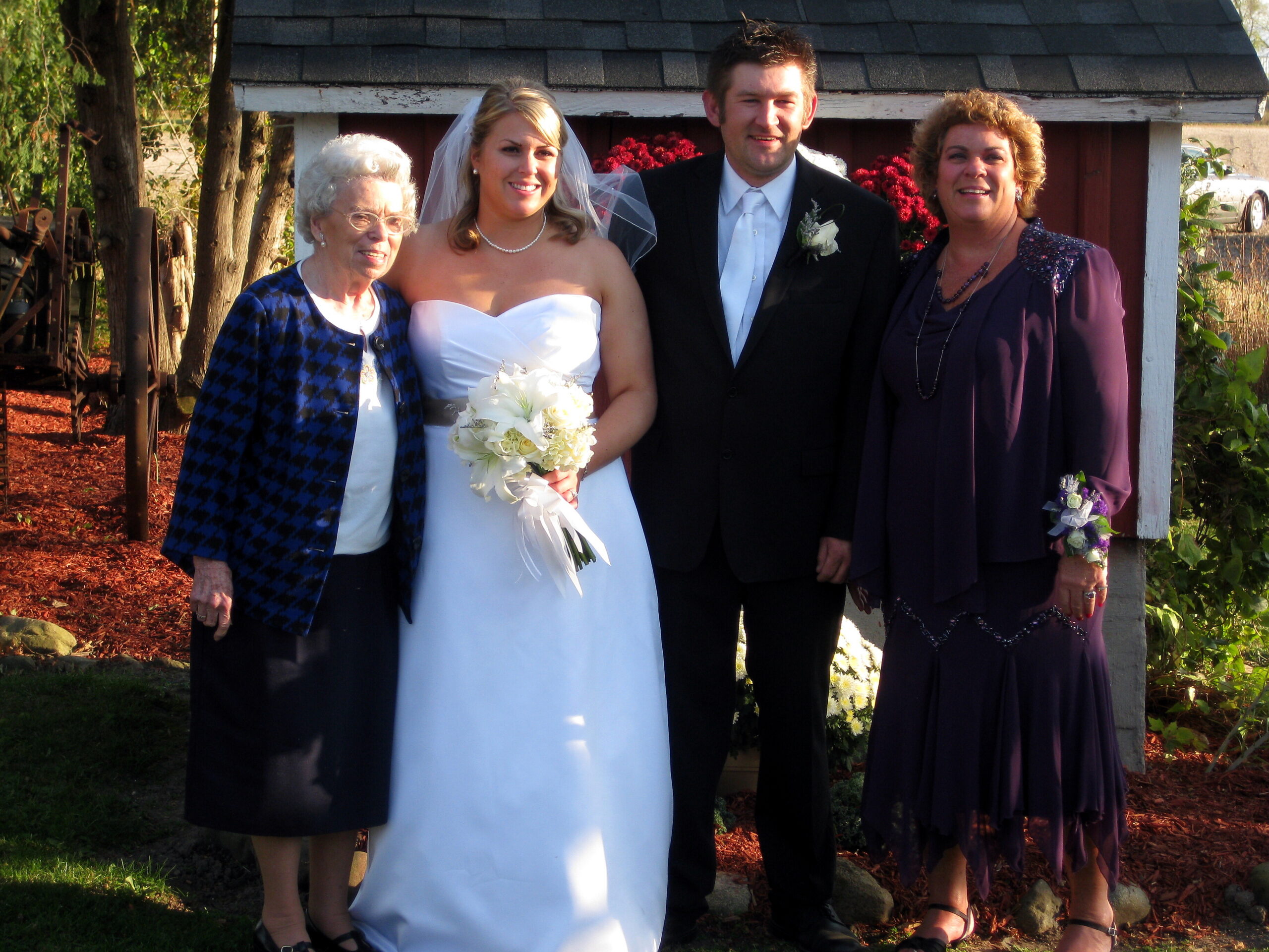 Bride in a white strapless gown holding a bouquet, flanked by groom in a suit and two older women outdoors at a wedding venue.