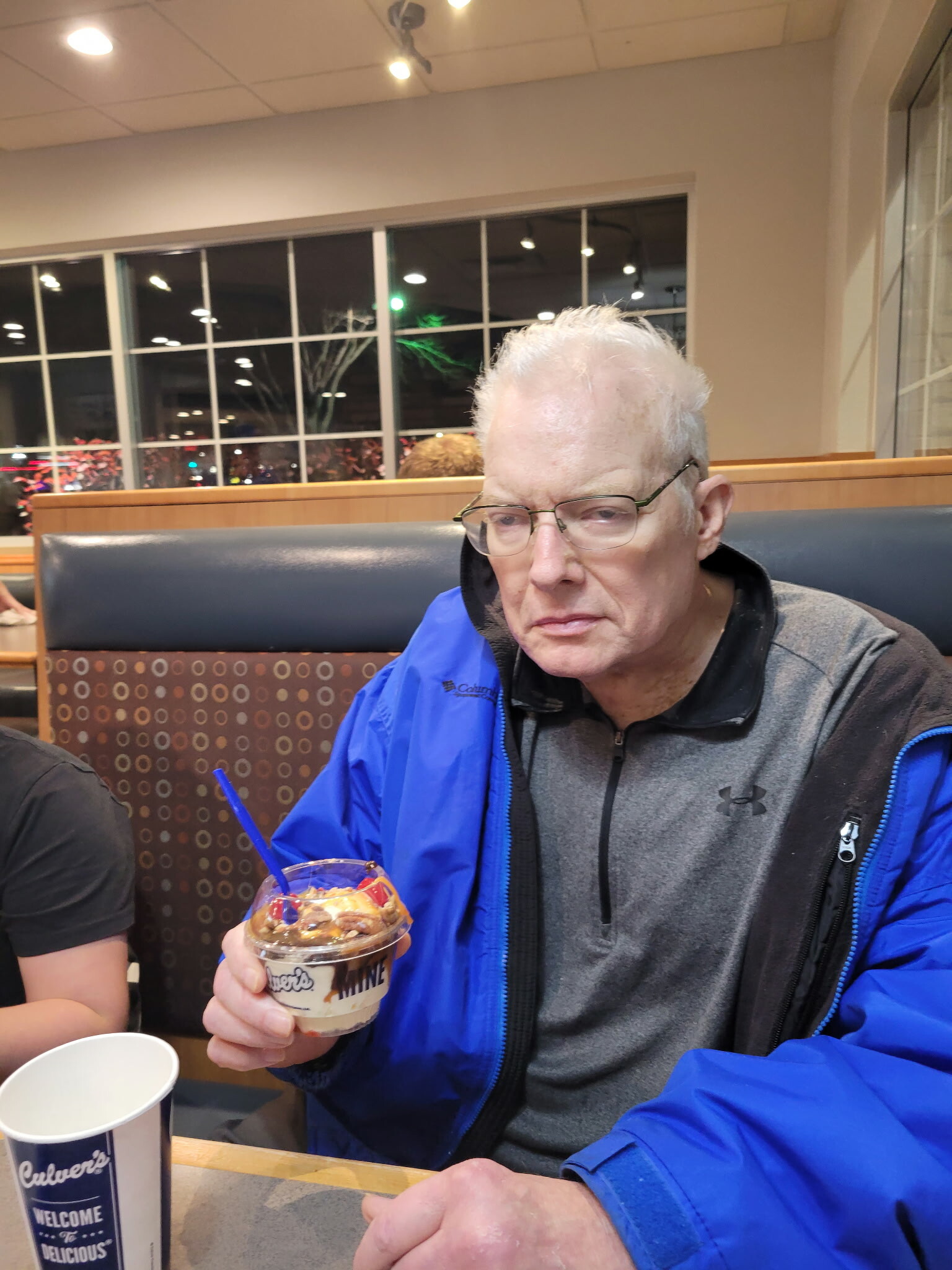 Elderly man with white hair and glasses in a blue jacket sits in a restaurant booth, holding a dessert cup with a blue straw.
