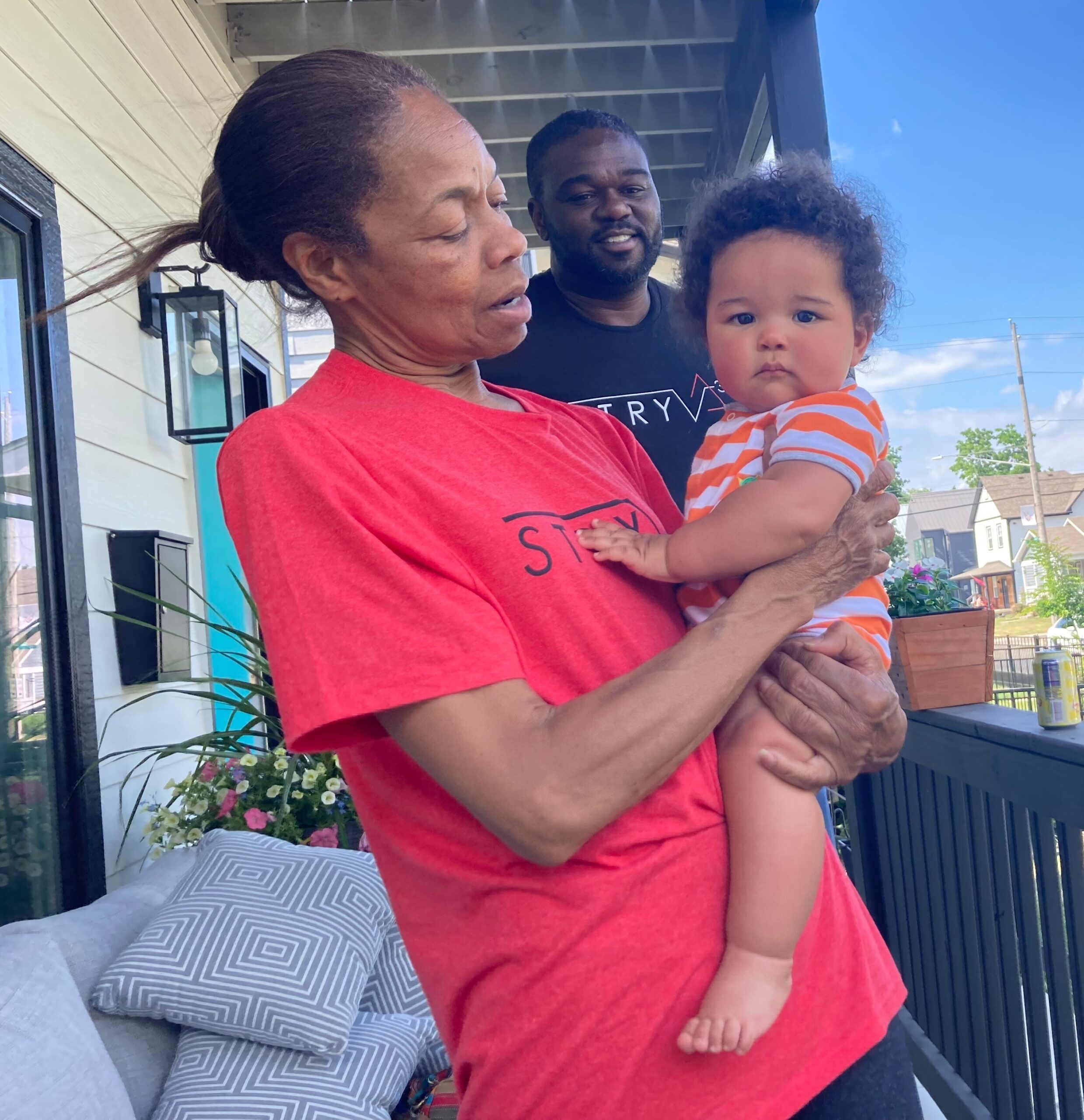 A man in a red shirt holds a baby in orange-and-white stripes outdoors on a porch, with another man in a black shirt smiling in the background.
