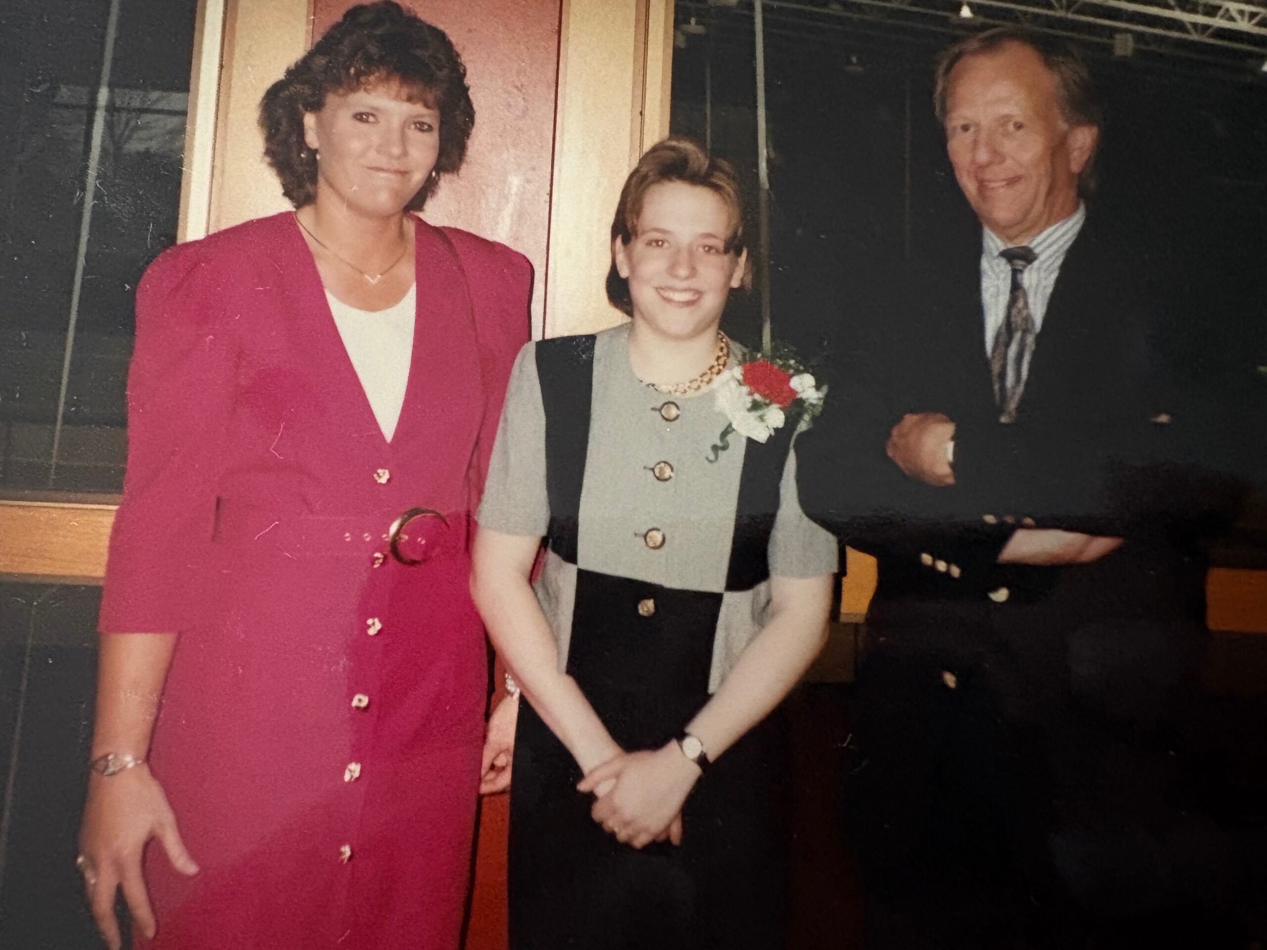 Three people pose for a photo indoors: woman in a magenta dress on the left, a young woman in gray with a corsage in the center, and a man in a suit on the right.
