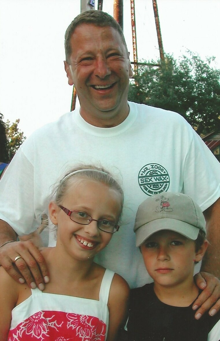 Smiling man with two children outdoors, arm around them, posing for a photo at what looks like an amusement park or fairground.