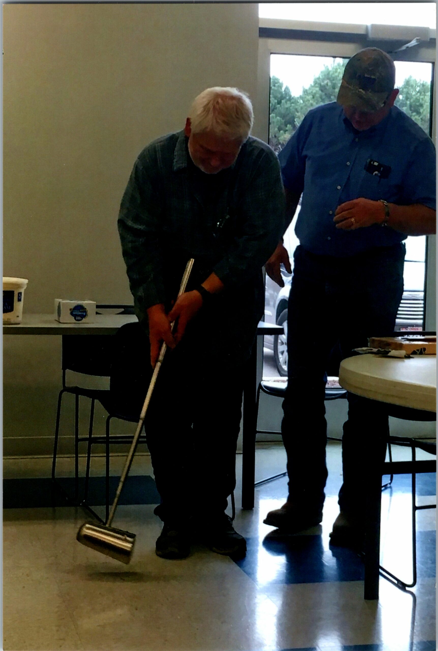 Two older men indoors, one bending over to handle a long metal tool while the other watches, in a room with a table and boxes in the background.