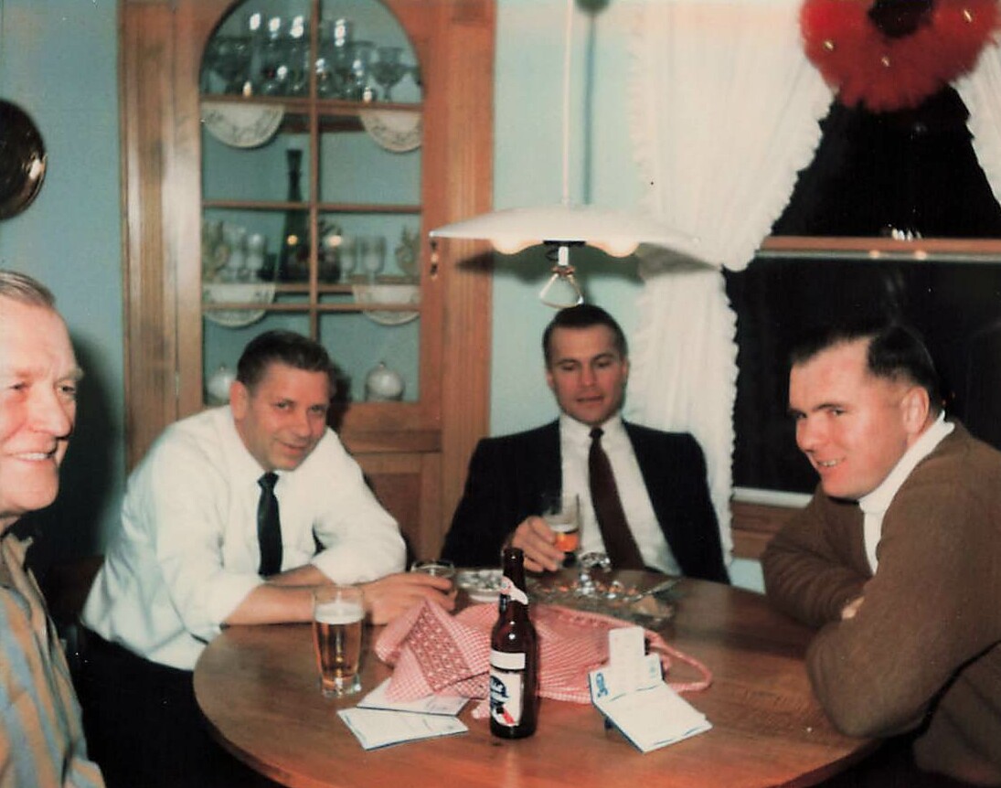 Four men sit around a round wooden table with beer and playing cards in a cozy dining room.