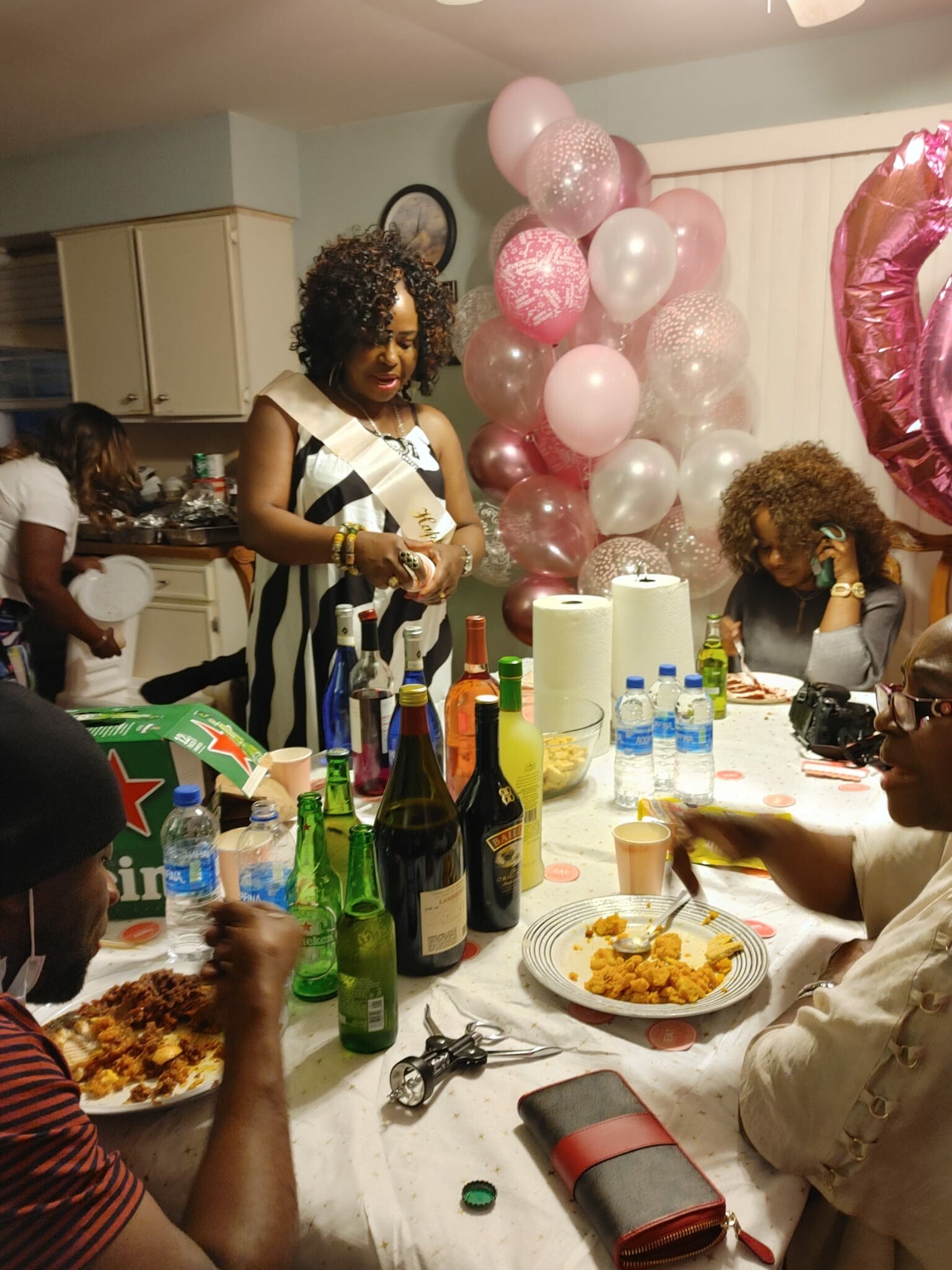 Woman wearing a sash standing by a table of drinks and snacks at a birthday party, pink balloon decorations in the background.
