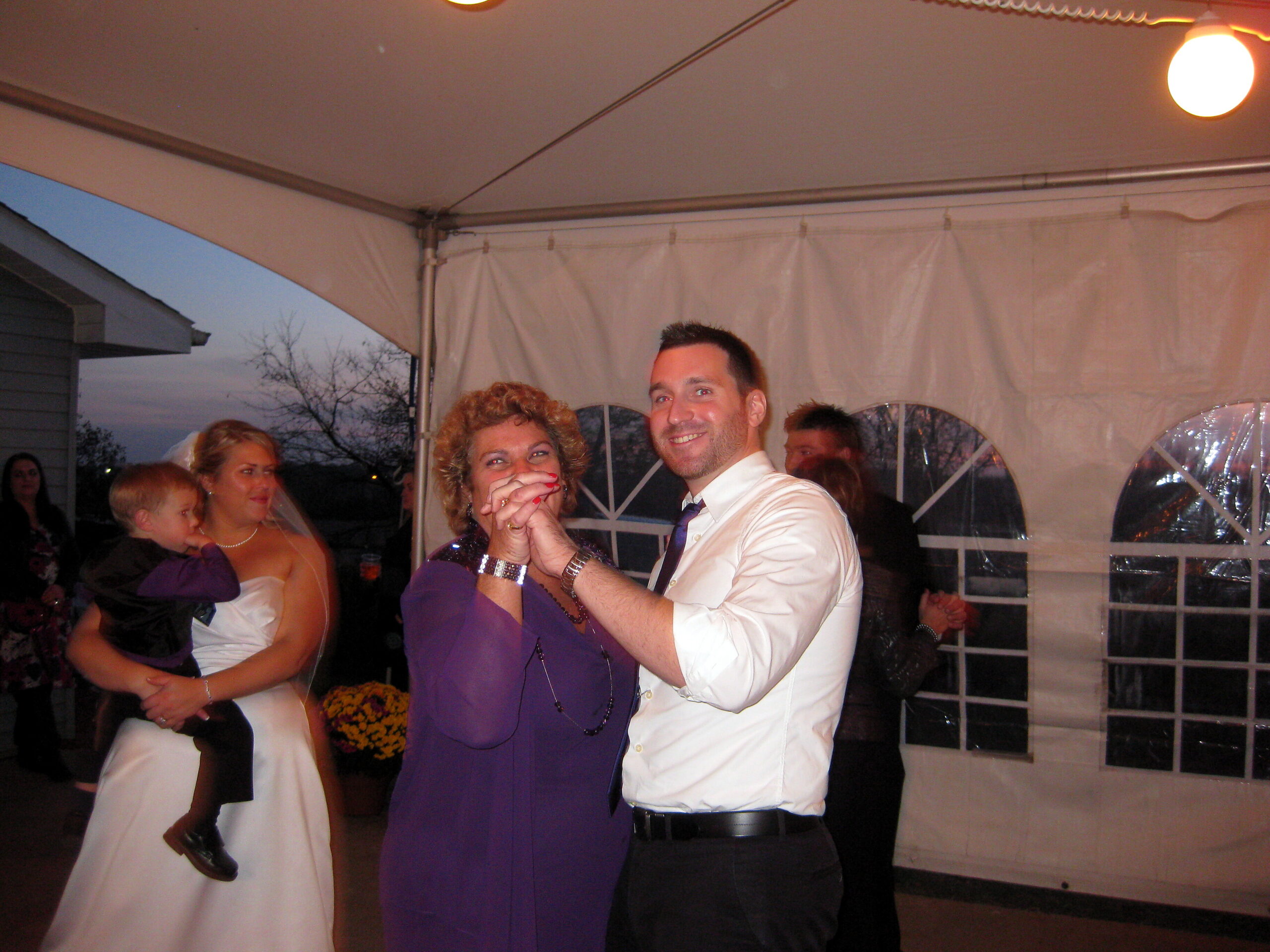 Wedding reception scene: couple dancing in a tent with guests watching in the background (woman in purple dress, man in white shirt).