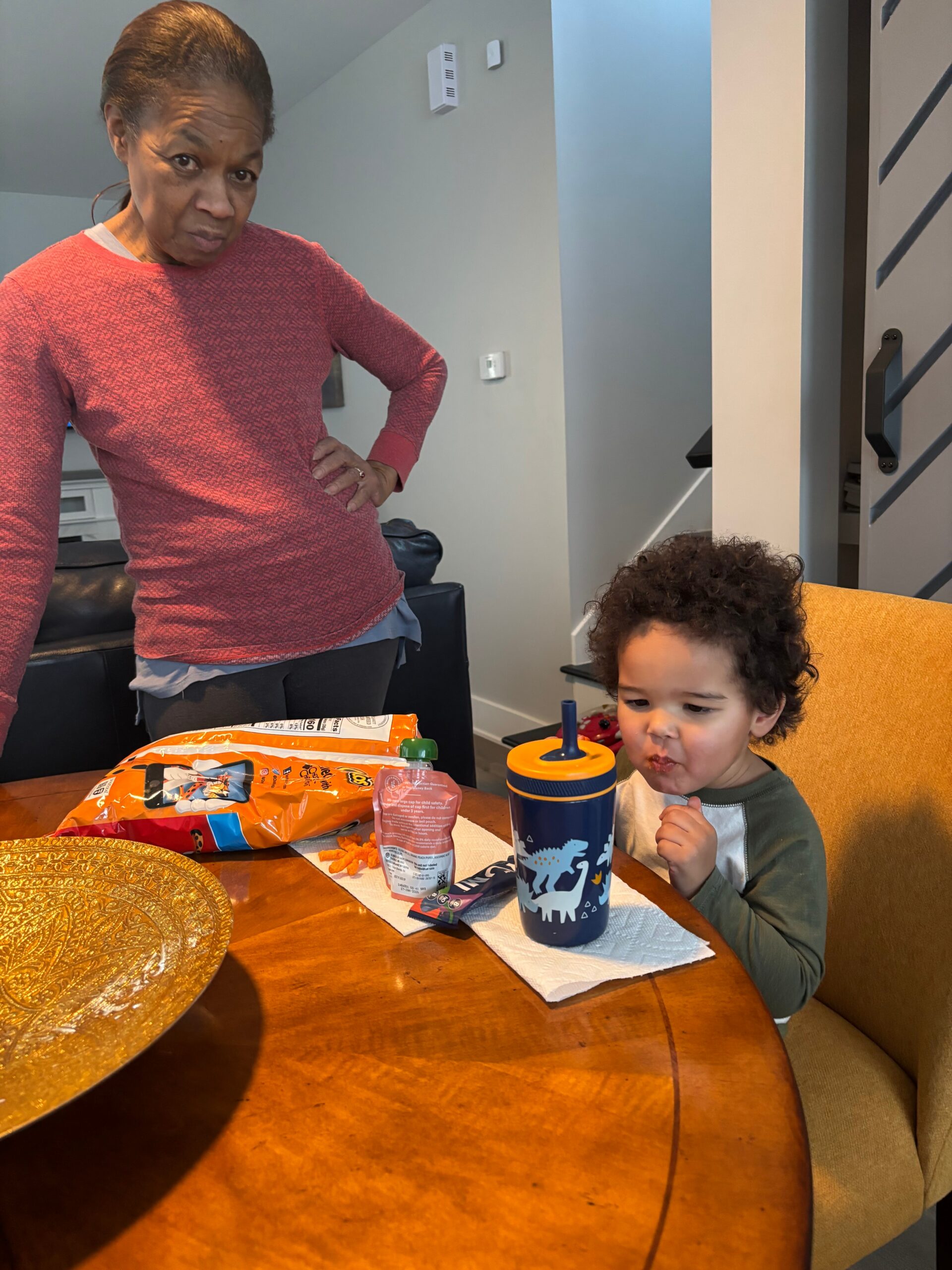 Woman standing with hand on hip beside a child at a dining table cluttered with snack bags and a dinosaur-themed sippy cup on a bright wooden table.