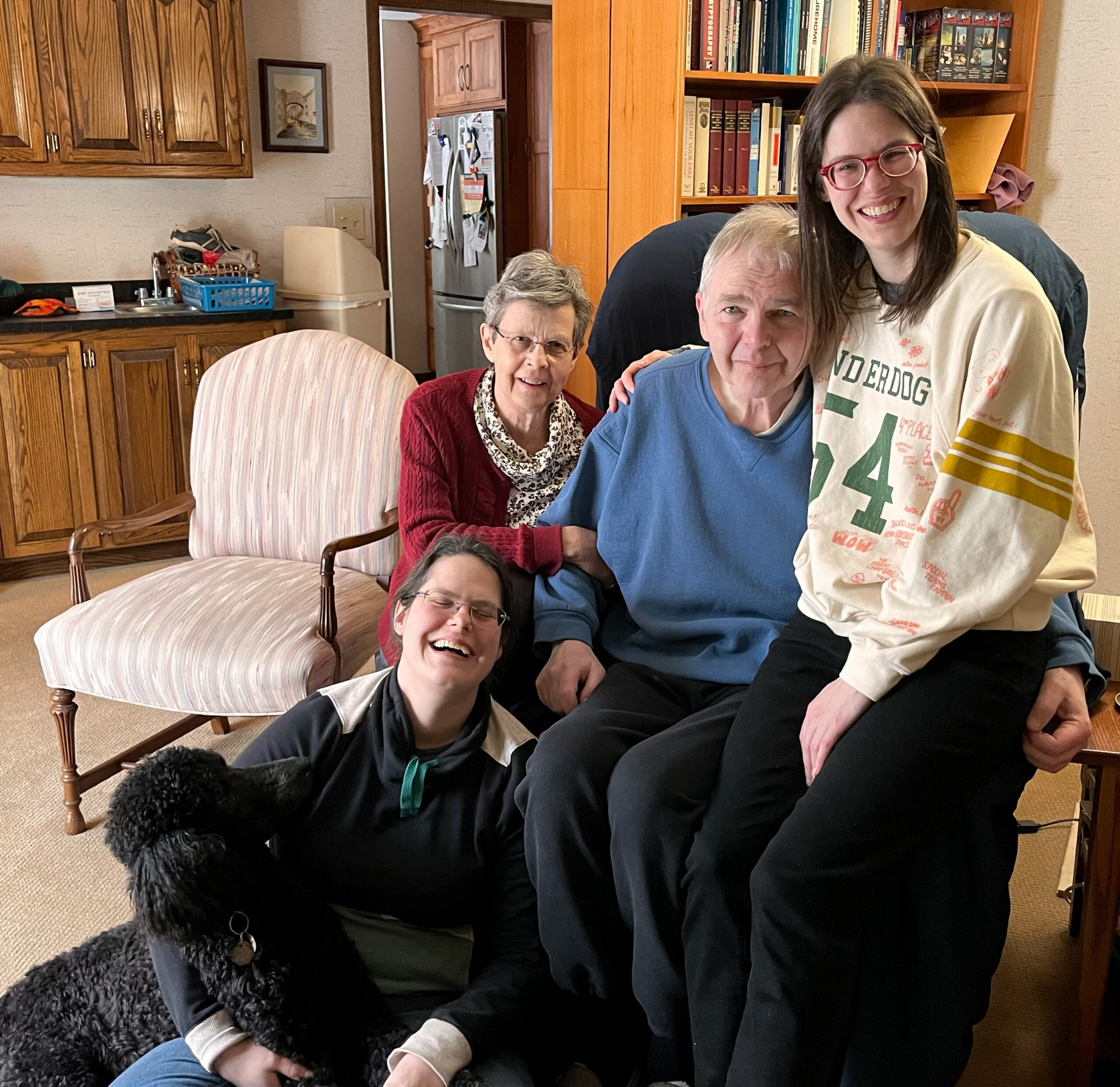 Smiling group in a living room with a fluffy black dog in the foreground and people seated together behind them. Bookshelf and kitchen area visible in the background.