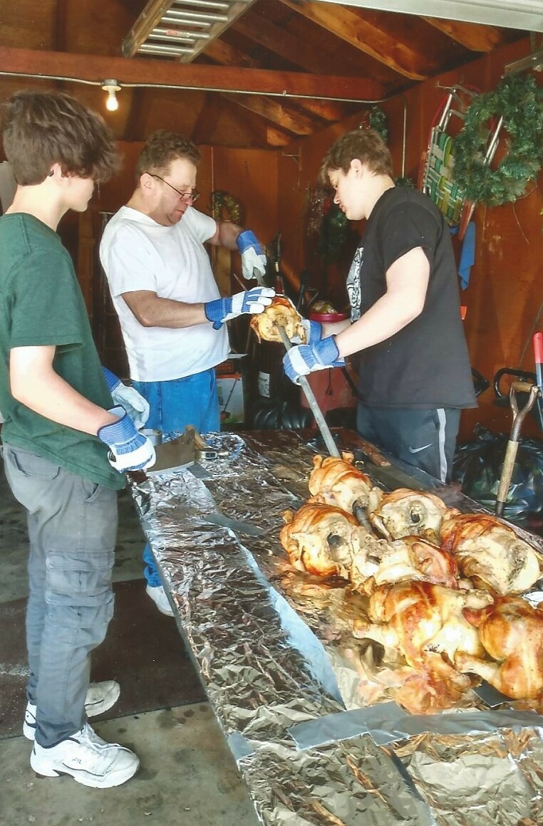 Three people wearing gloves carve roasted meat on a foil-covered table in a garage or workshop, with one person pulling meat from a roaster while others assist.