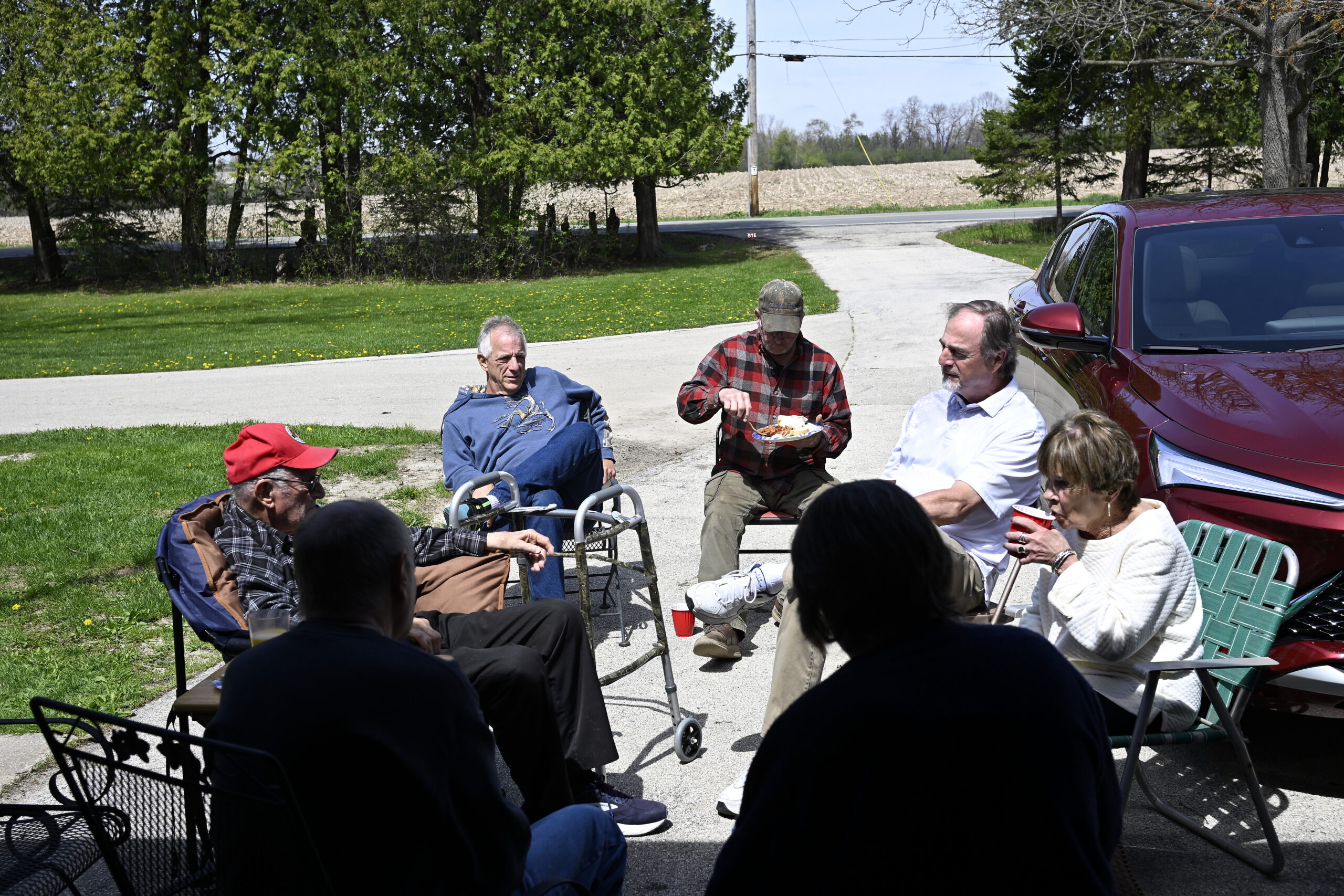 Group of seniors seated in lawn chairs and a walker in a driveway, sharing food and drinks on a sunny day near a red SUV.