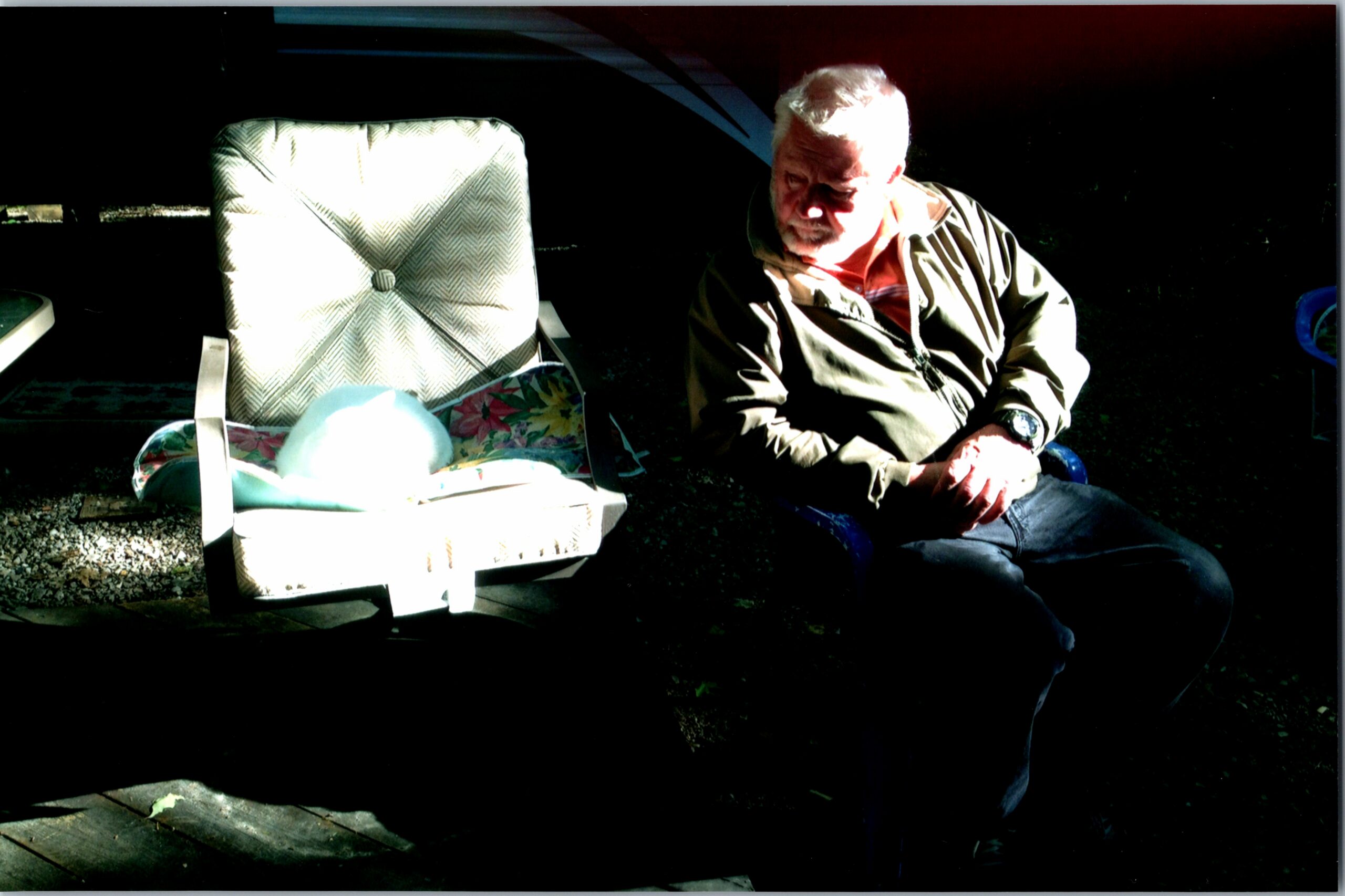 Older man sitting on the ground beside a patterned outdoor chair at night, looking downward.