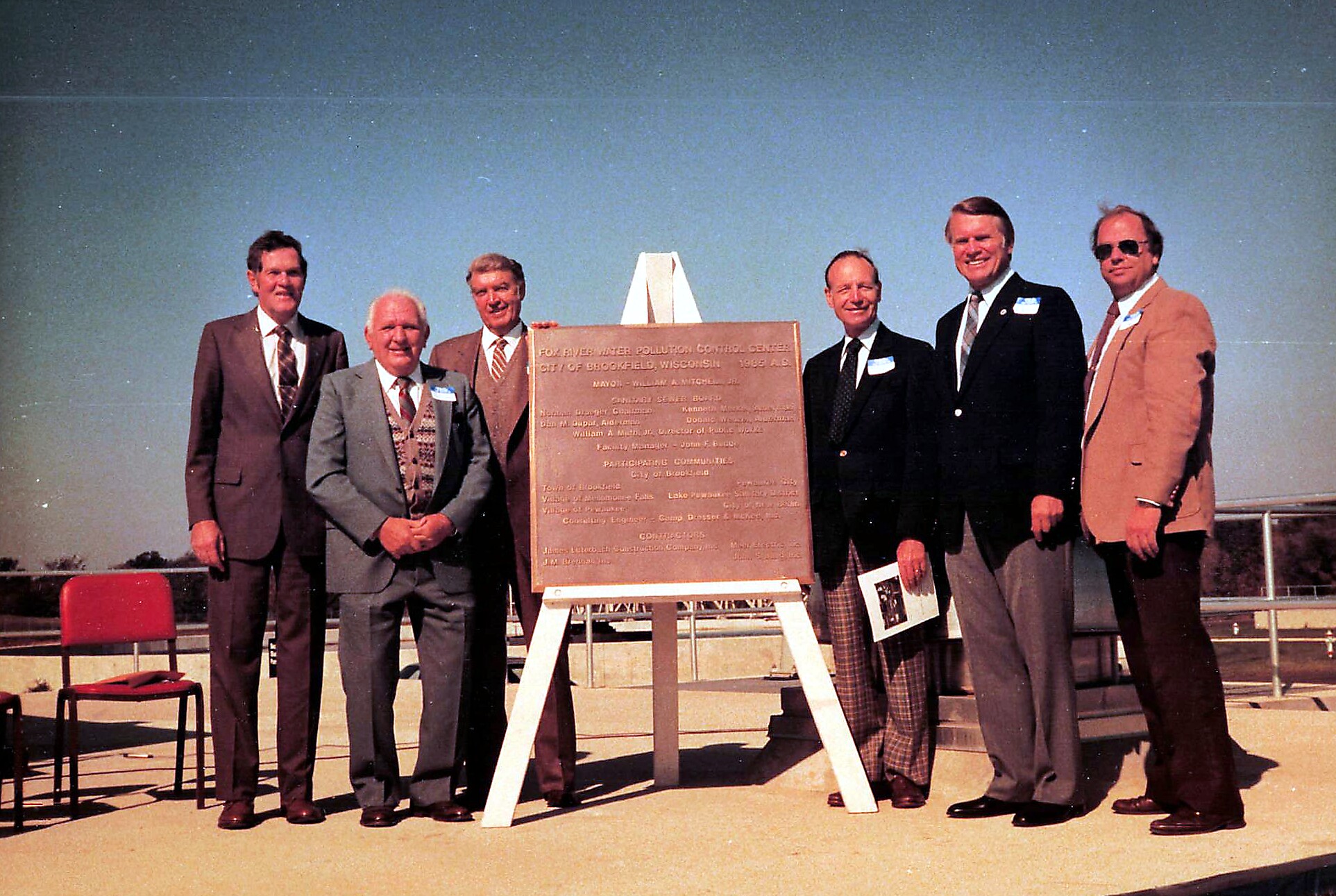 Six men in suits stand around a wooden plaque on a white easel outdoors, with a clear blue sky behind them and red chairs nearby.