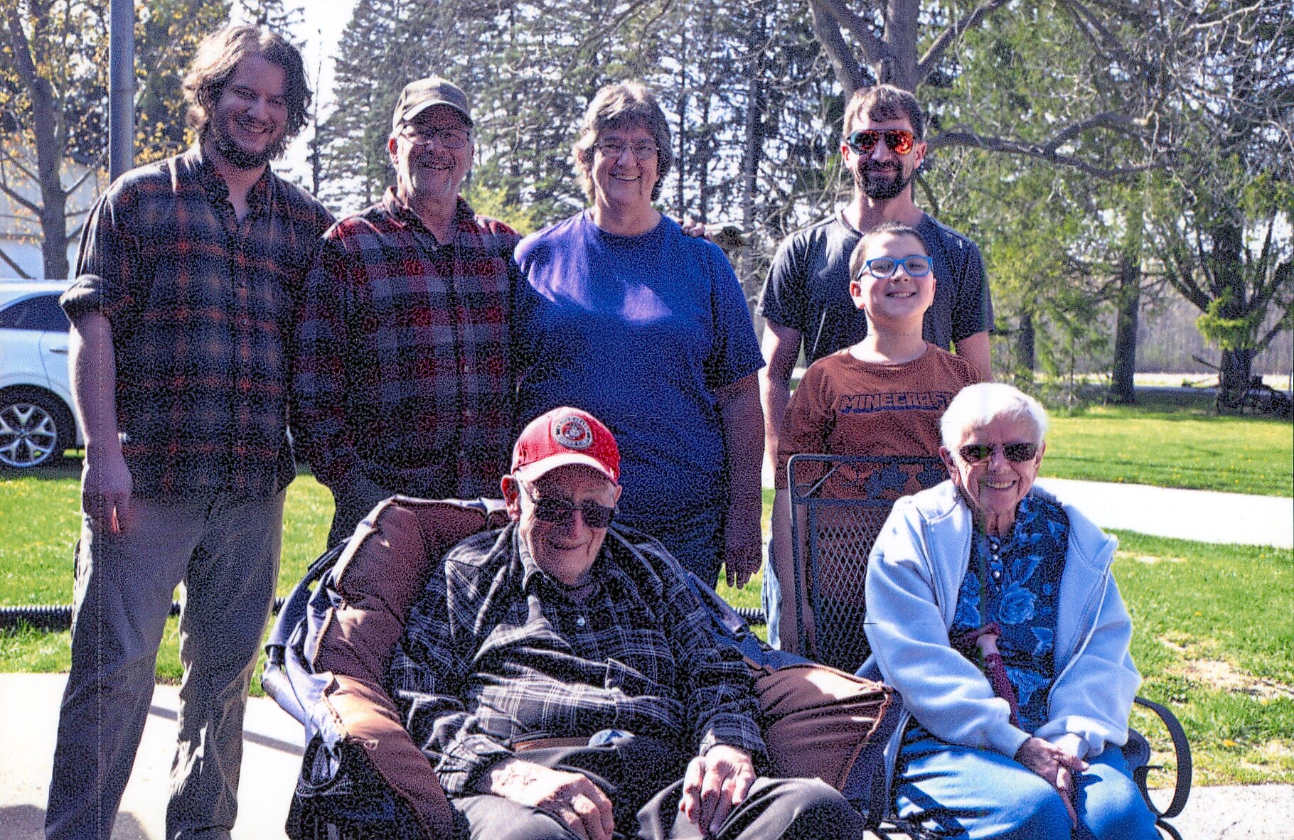 Group of seven people posing outdoors on a sunny day, two seniors in wheelchairs in front and five adults standing behind, smiles all around in a park-like setting.