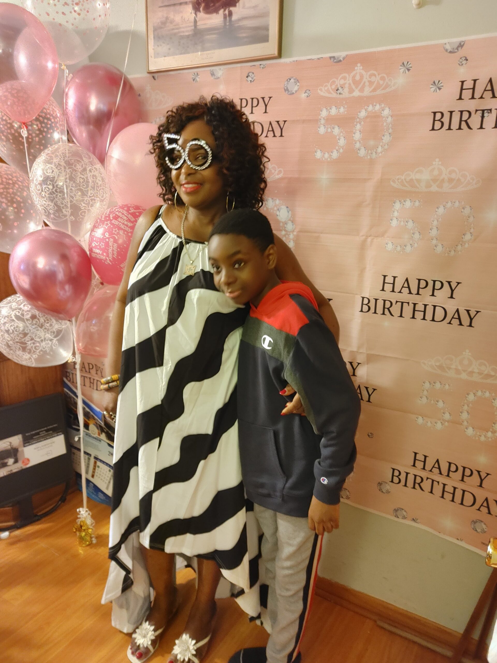 Woman in a black-and-white zebra-print dress wearing oversized '50' glasses poses with a boy at a pink birthday backdrop with balloons.