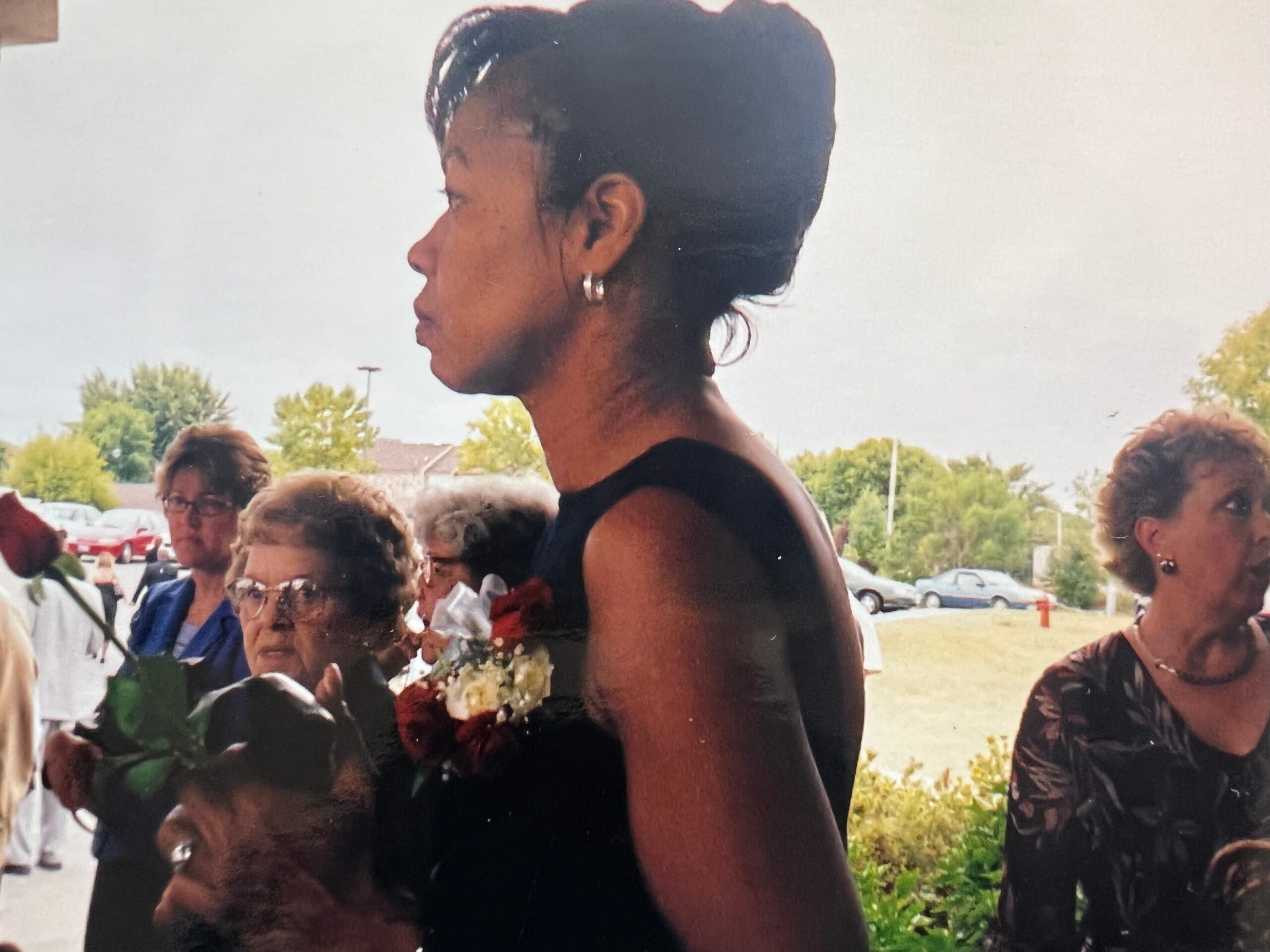 A woman in a black sleeveless dress seen in profile at an outdoor event, wearing a flower garland around her neck as attendees stand nearby.
