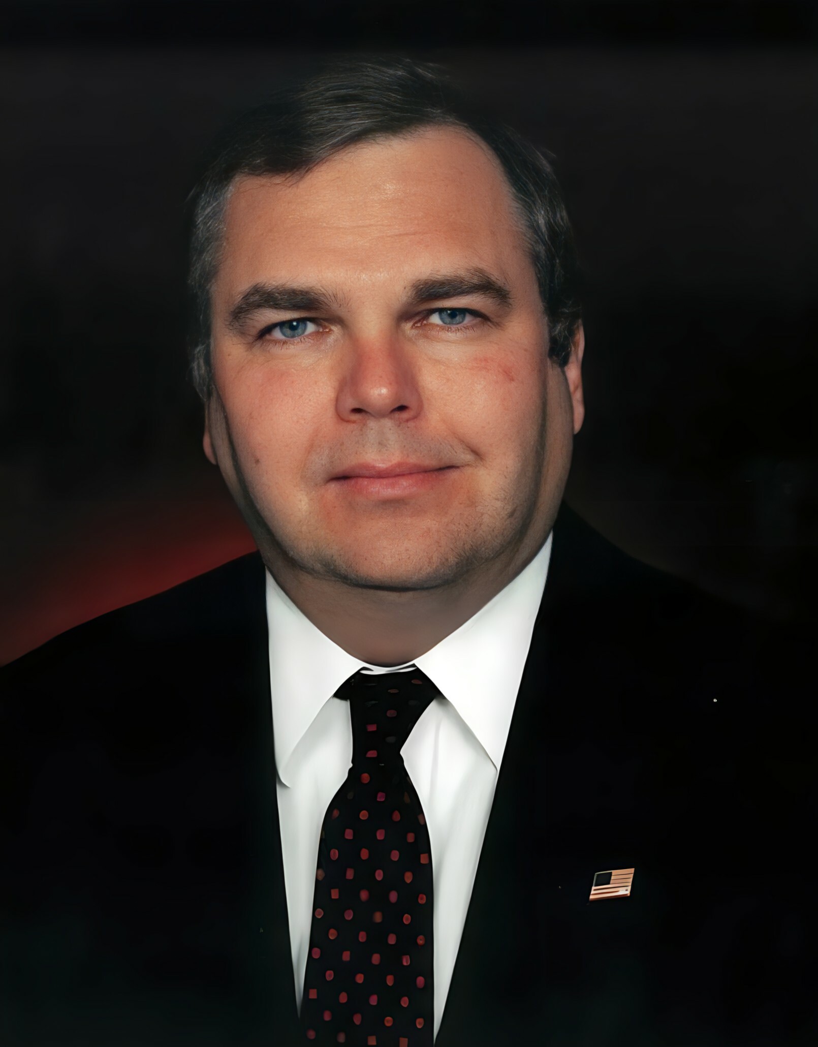 Close-up portrait of a man in a black suit and red dotted tie against a dark background, looking at the camera.