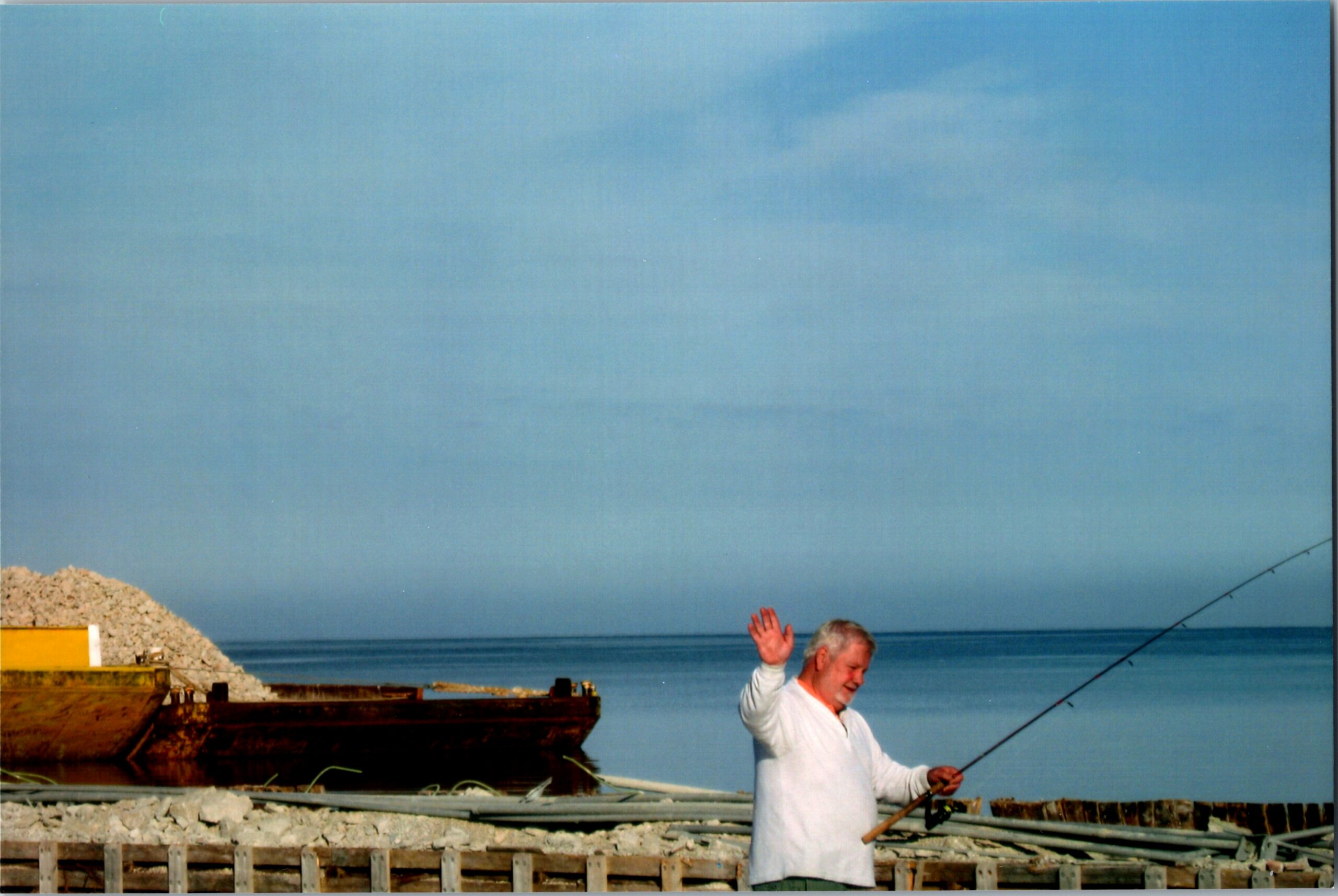 Man in white shirt near a rocky shoreline at a construction site, raising his right hand as a fishing rod extends toward the water behind him