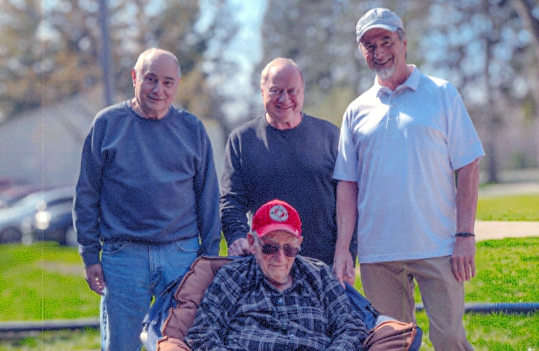 Four men outdoors posing; an elderly man in a wheelchair with a red cap sits in front while three others stand behind on a sunny day.
