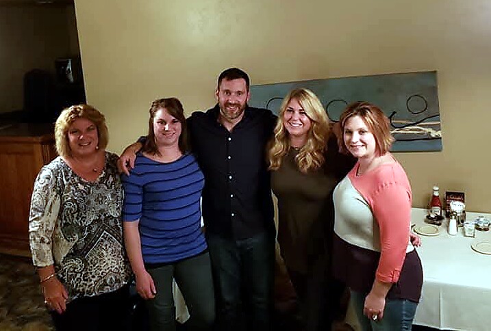 Five adults posing together in a restaurant, smiling at the camera; a man in a black shirt stands center with four women around him on a beige wall backdrop.