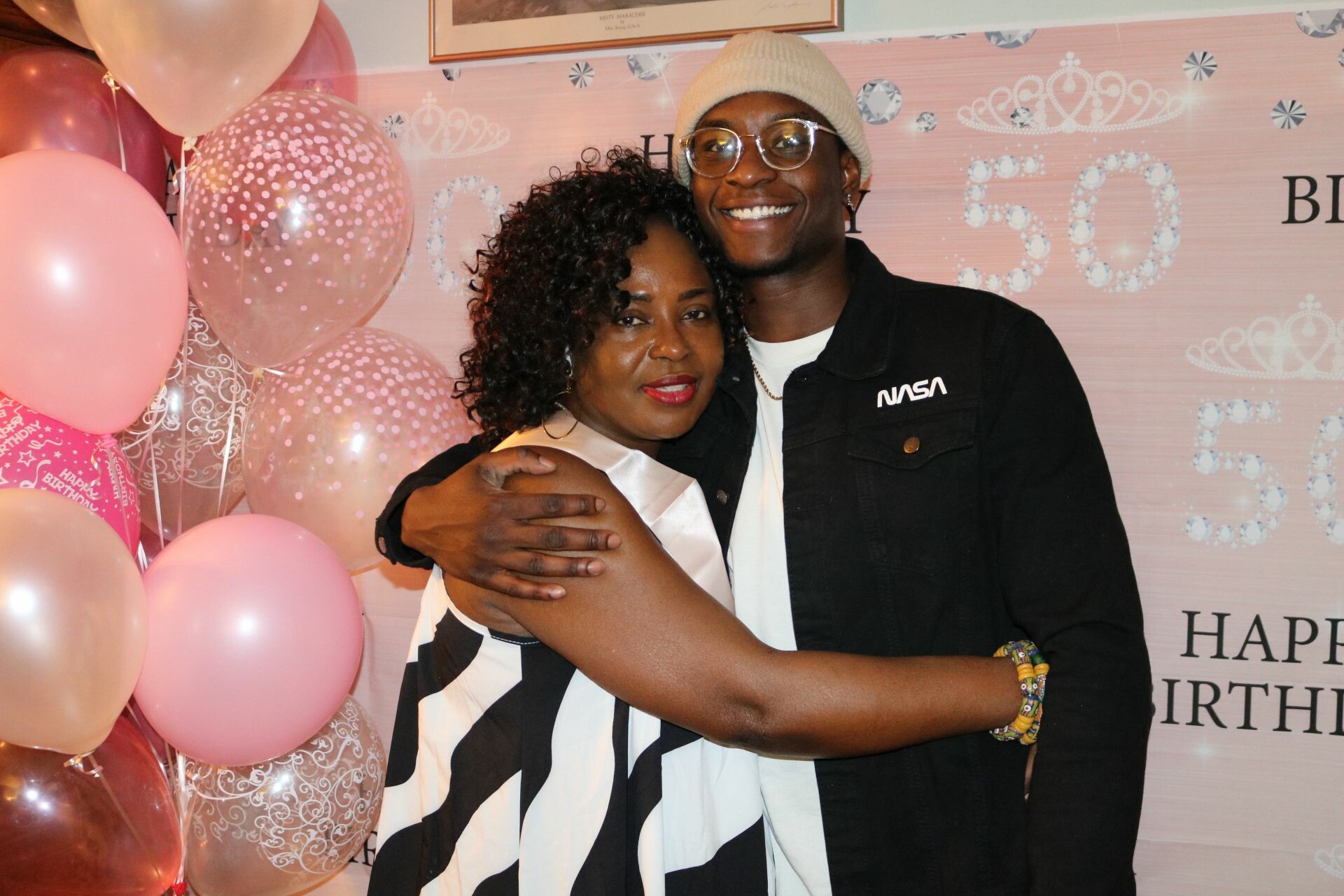 Couple hugging and smiling at a birthday party with pink balloons and a decorative backdrop saying Happy Birthday
