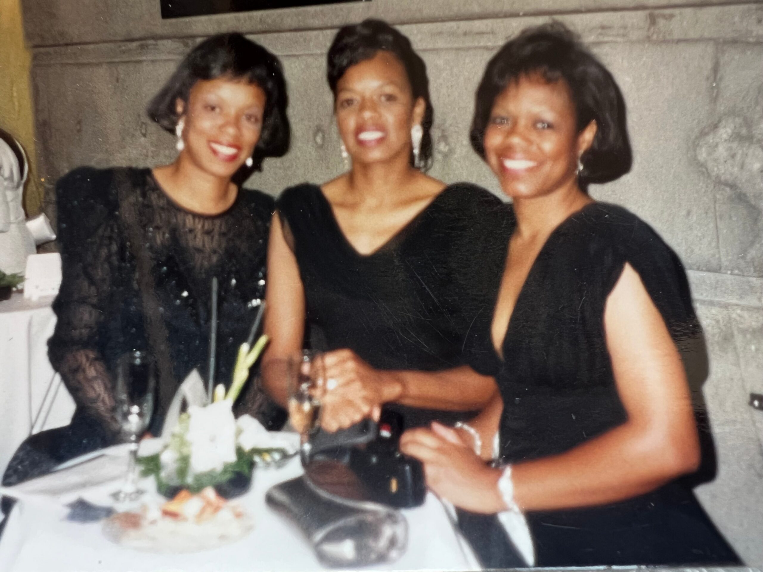 Three women in black dresses smile at a celebratory table with a cake and flowers in front of them, posing for a photo.