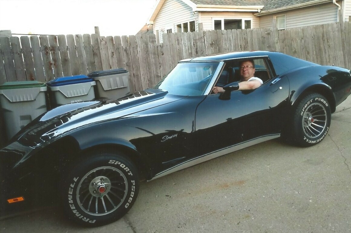 A man sits in the driver’s seat of a shiny black classic Chevrolet Corvette in a residential driveway, with a wooden fence and trash bins nearby.