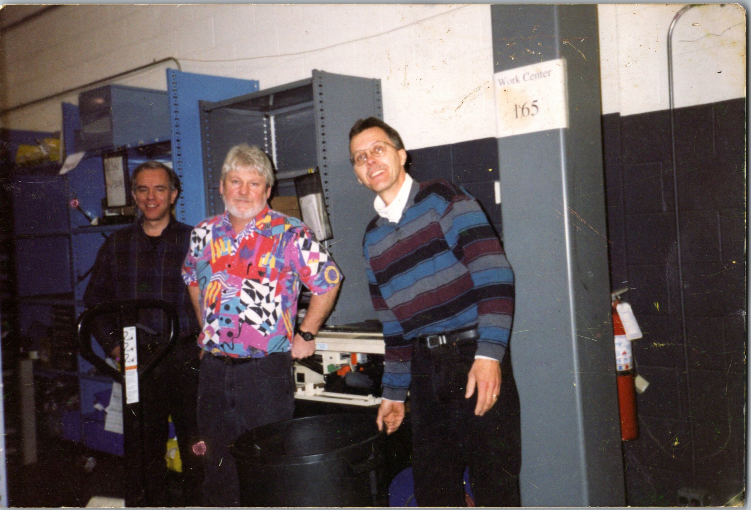 Three men in a workshop posing beside blue industrial cabinets; sign reads Work Center 165