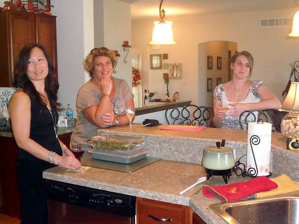 Three women stand at a kitchen island, each holding a drink and smiling at the camera.