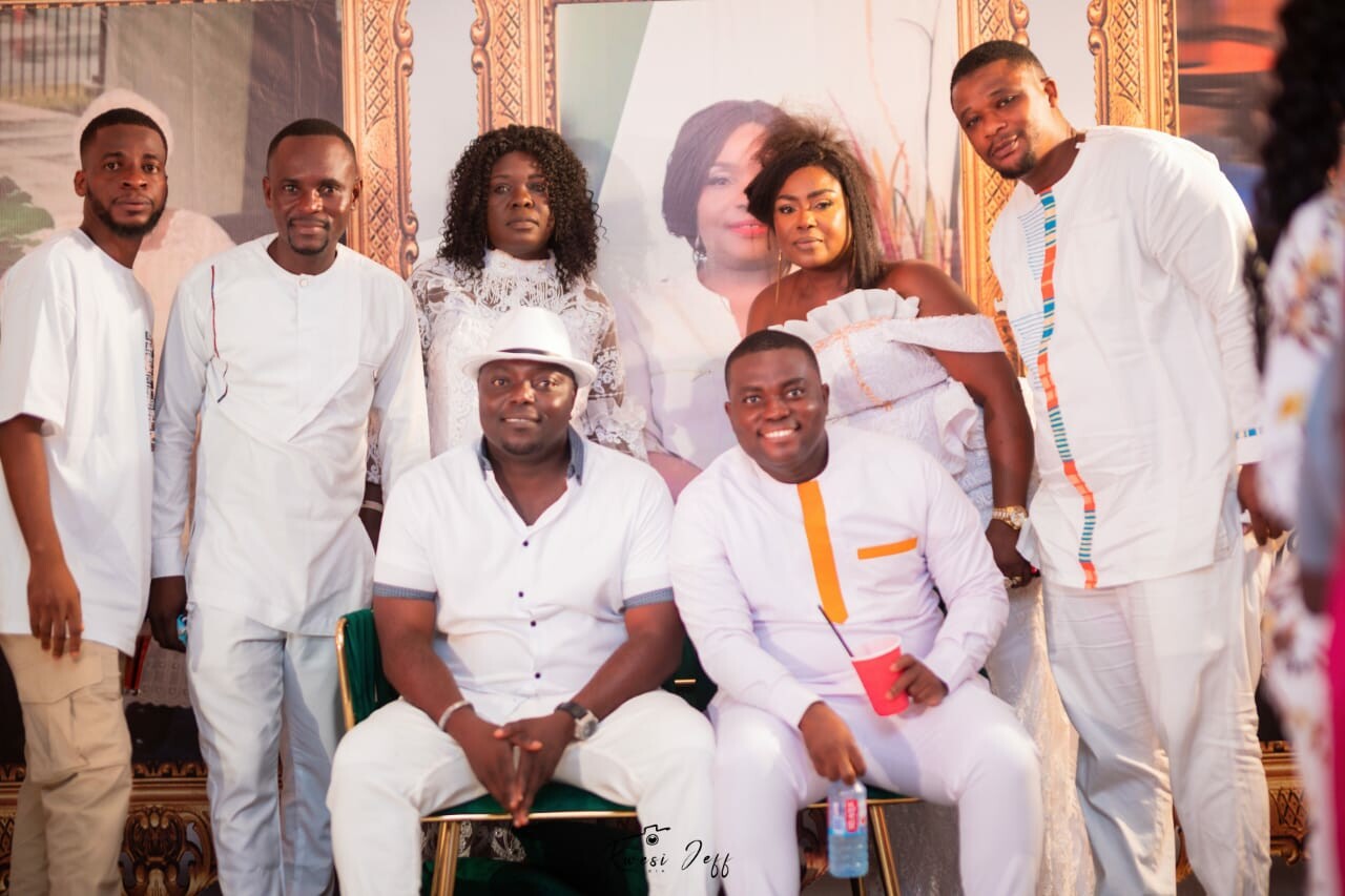 Group portrait of eight people in white outfits posing in front of an ornate backdrop at a celebration, two men seated center with drinks nearby.