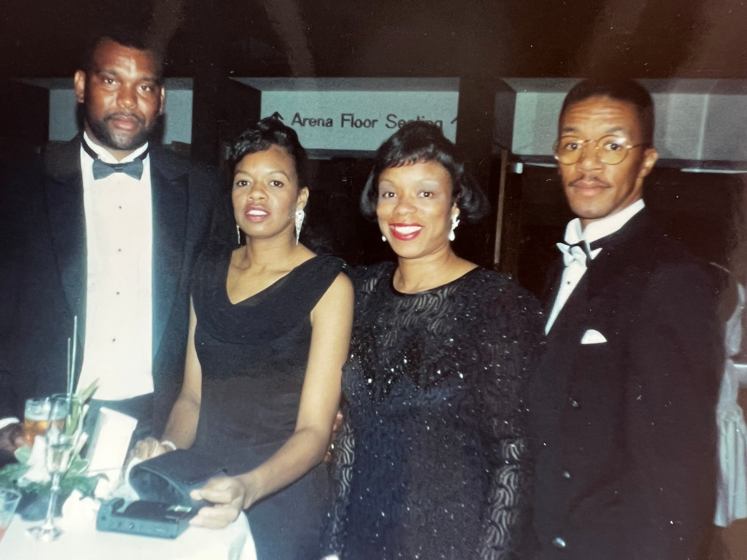 Four adults in formal attire posing for a photo at a black-tie event; two men in tuxedos flanking two women in black dresses, standing behind a table with a laptop and a glass visible at the edge of the frame.