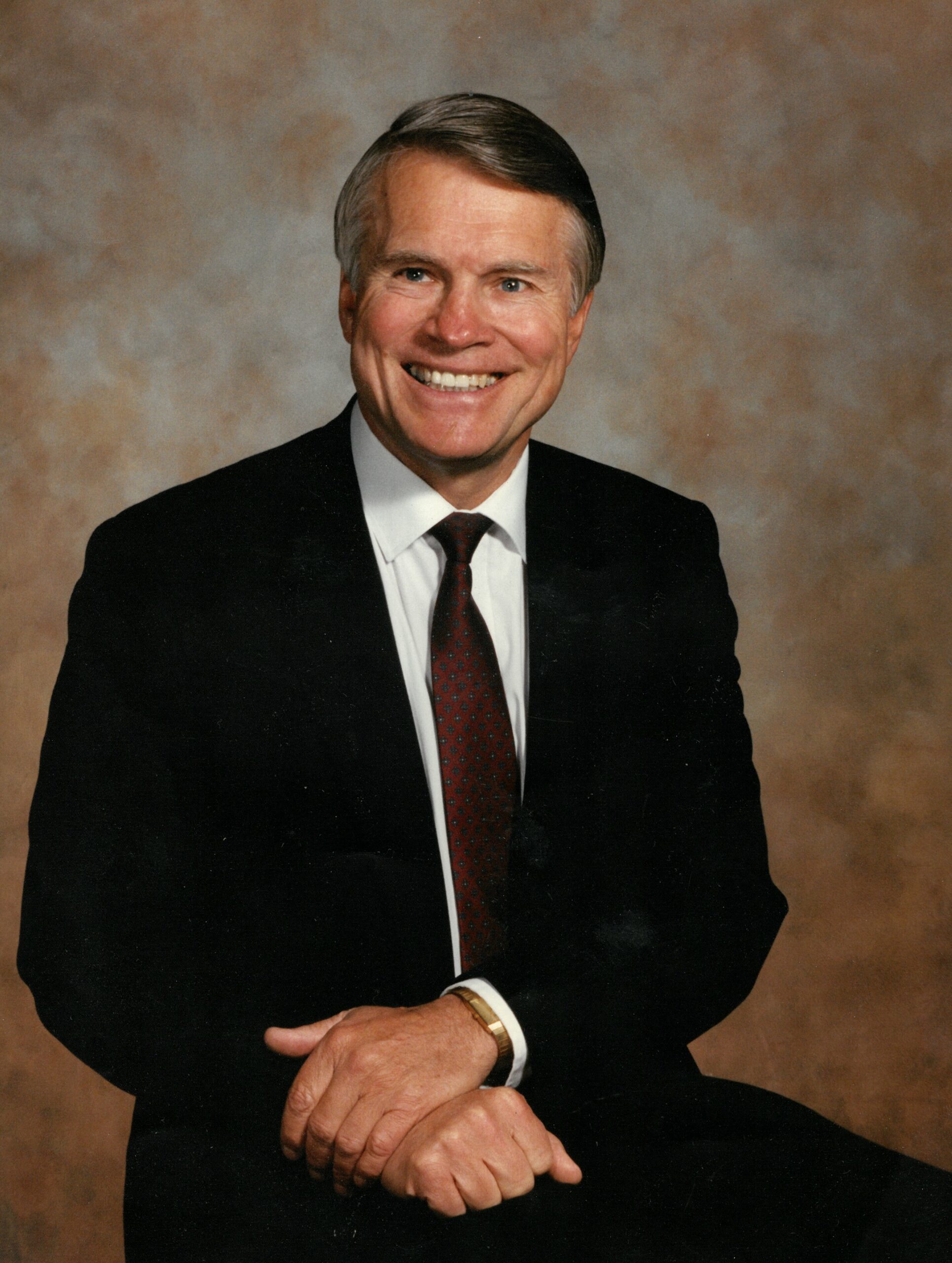 Portrait of a smiling older man in a dark suit, white shirt, and red tie, with hands folded on his lap.