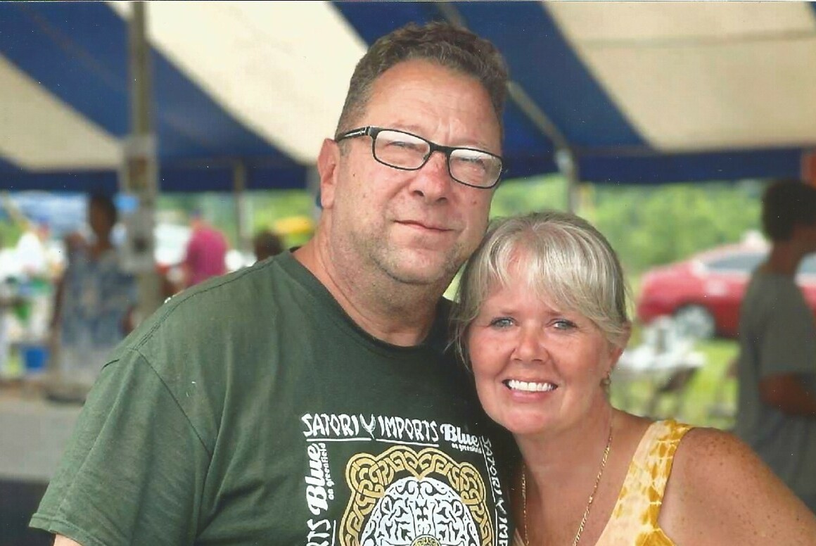 Smiling middle-aged couple posing together at an outdoor festival under a blue-and-white canopy.