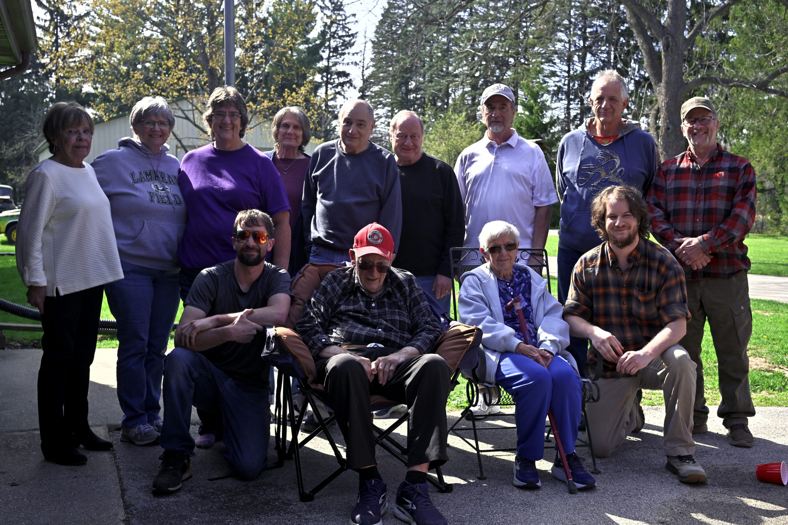 Group of about 14 adults outdoors posing for a photo in a sunny park, with trees in the background.