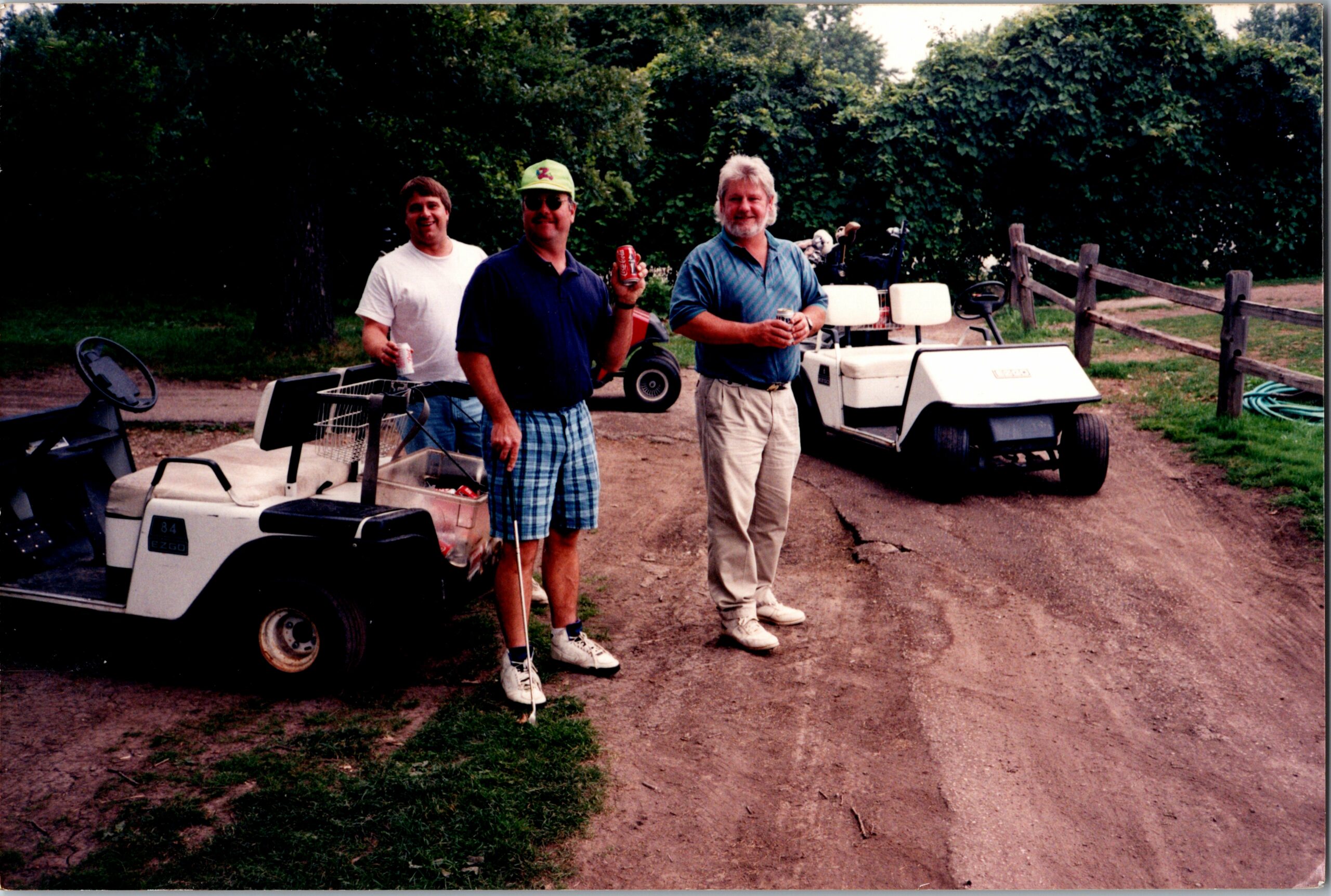 Three men stand outdoors beside golf carts on a dirt path, each holding a drink and smiling.