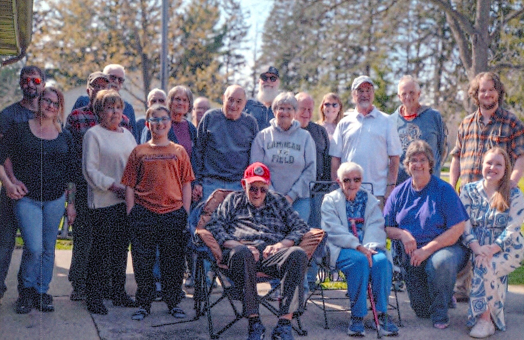 Group of adults posing outdoors for a group photo in a park, with two people seated in front and others standing behind them.