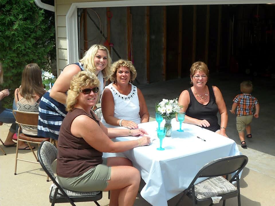 Four women sit around a table with a white tablecloth and turquoise drinks, smiling at a casual outdoor gathering in a driveway area near a garage.