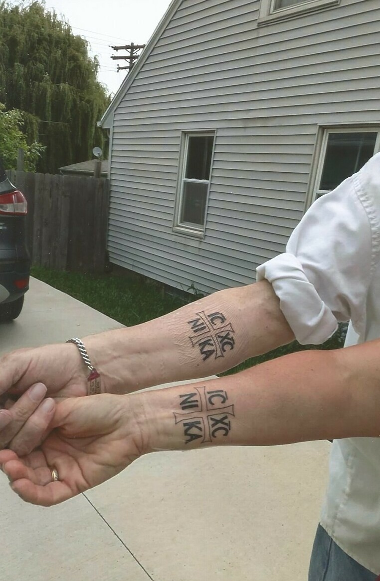 Close-up of two forearms extended outdoors, each with a black tattoo showing the Greek letters IC XC NIKA inside a cross, indicating a religious inscription.