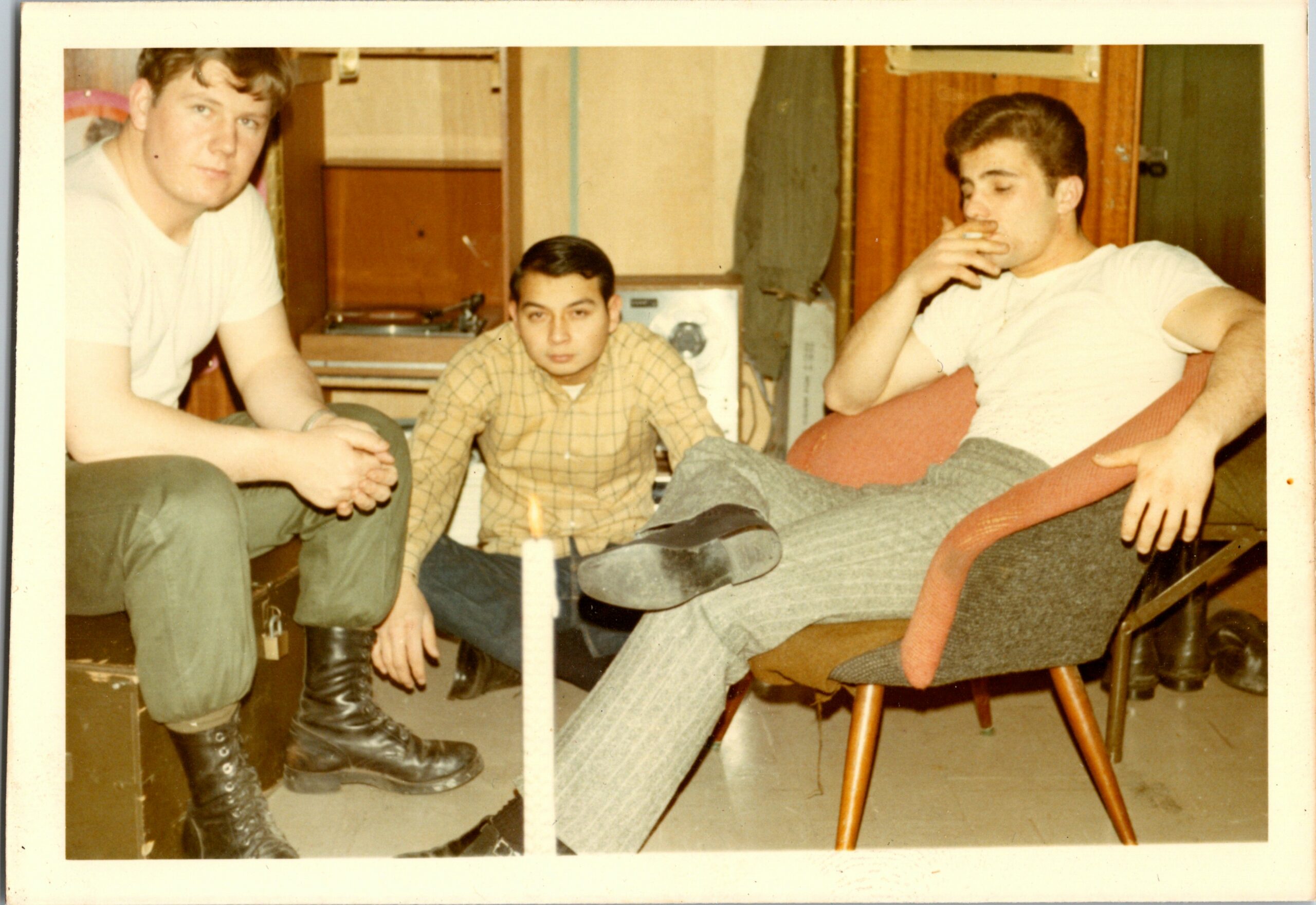 Three young men sit in a cluttered room, one wearing a white T‑shirt and boots, another in a plaid shirt, and the third lounging in a chair smoking a cigarette.