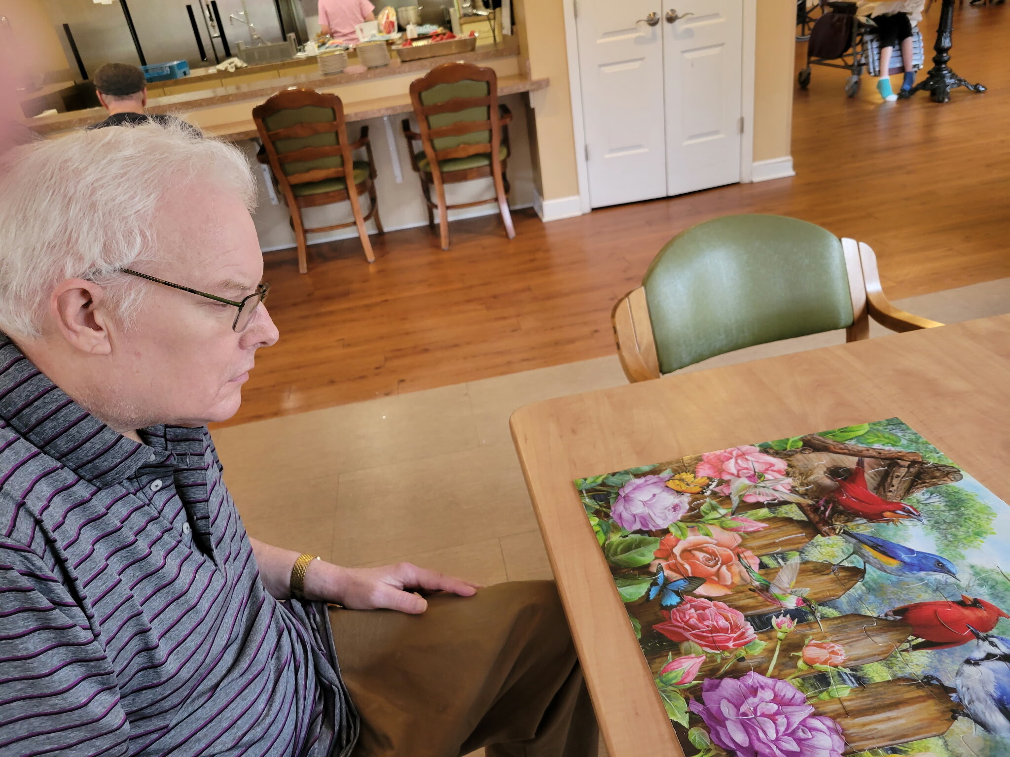 Close-up of an older man with white hair and glasses, seated at a table, looking down at a colorful floral puzzle spread before him.