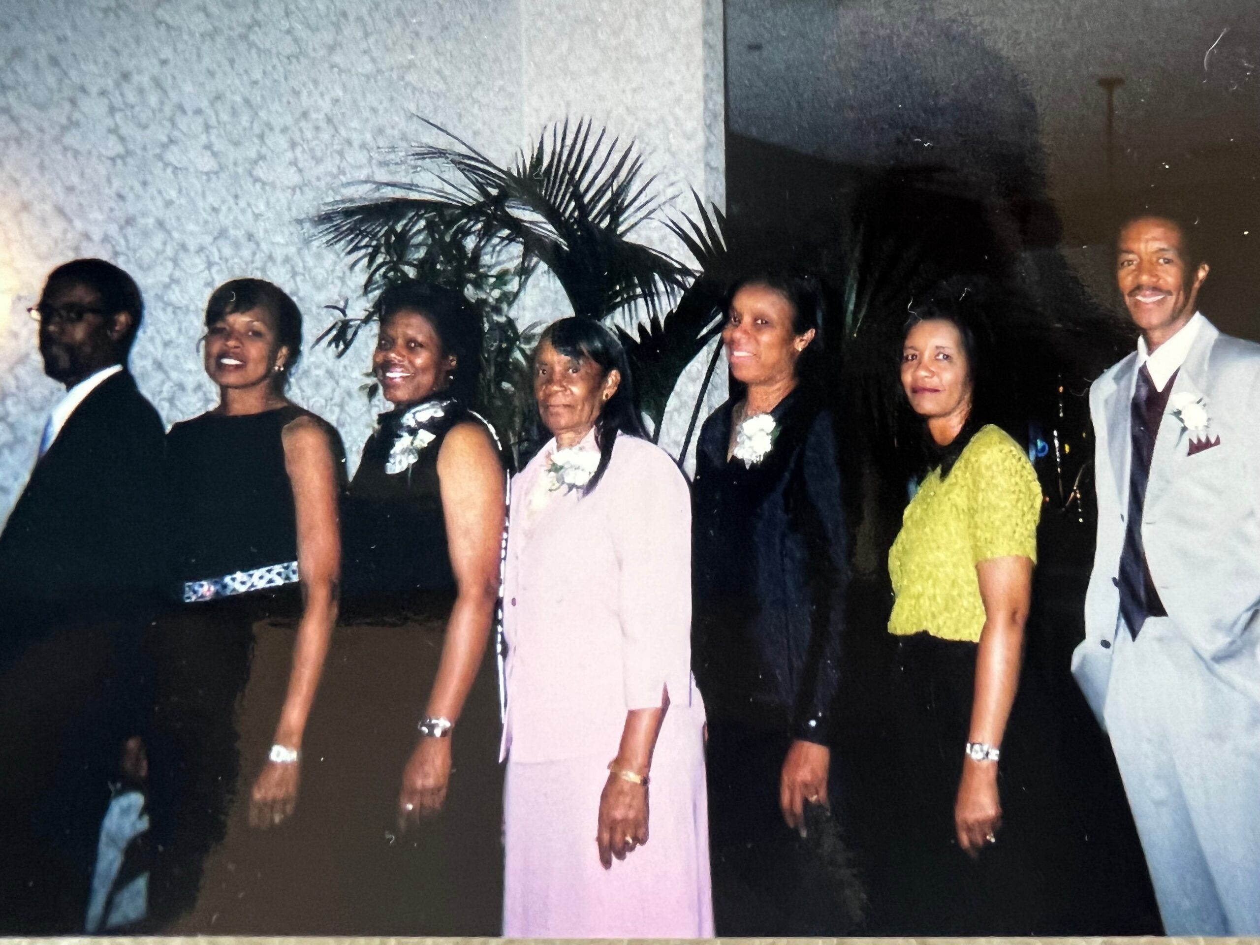 Group of adults in formal attire standing in a line for a posed photo, with a large plant and textured wall behind them.
