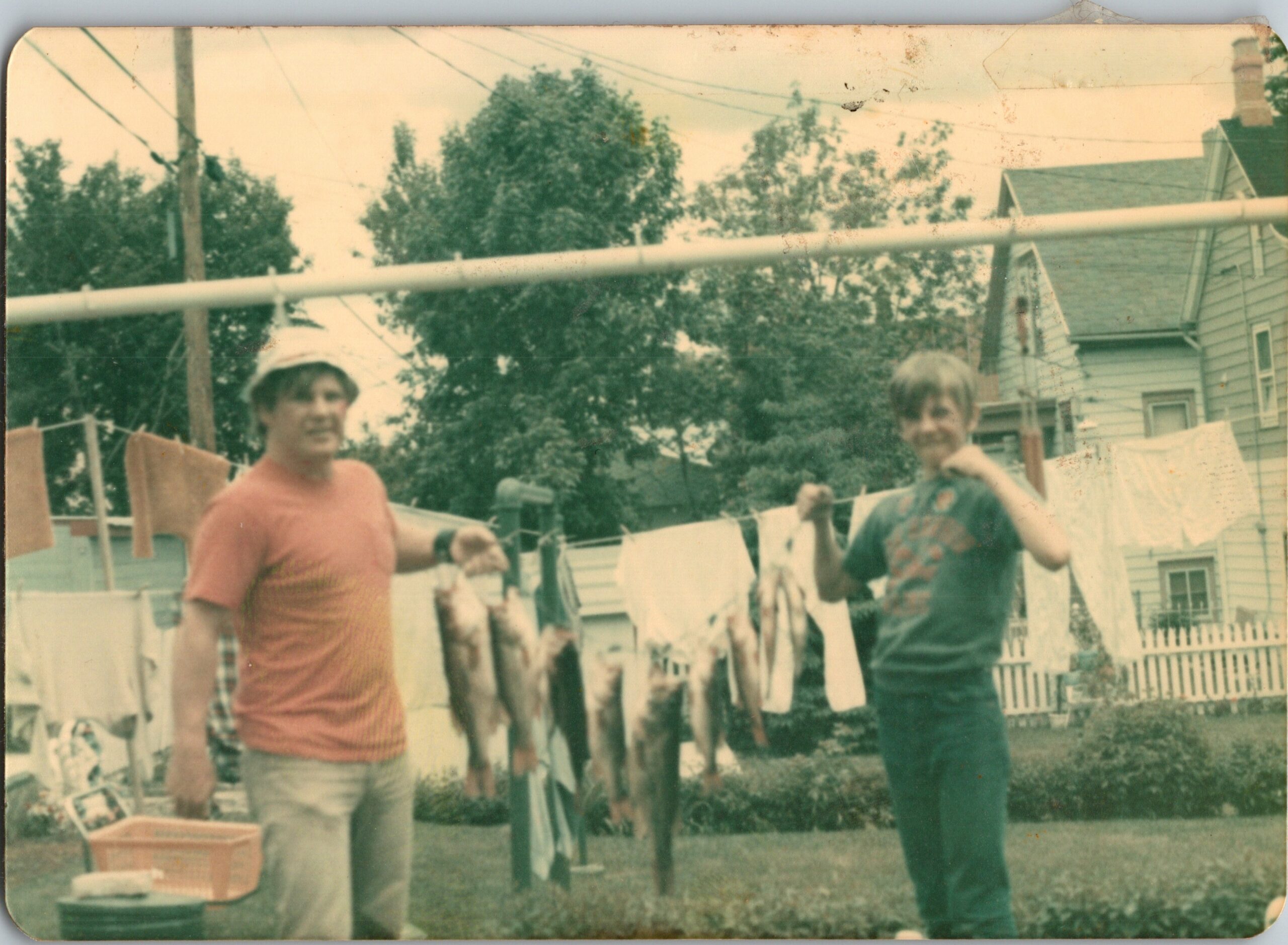 Two children stand beside a clothesline in a suburban backyard, with laundry hanging and houses in view behind them.