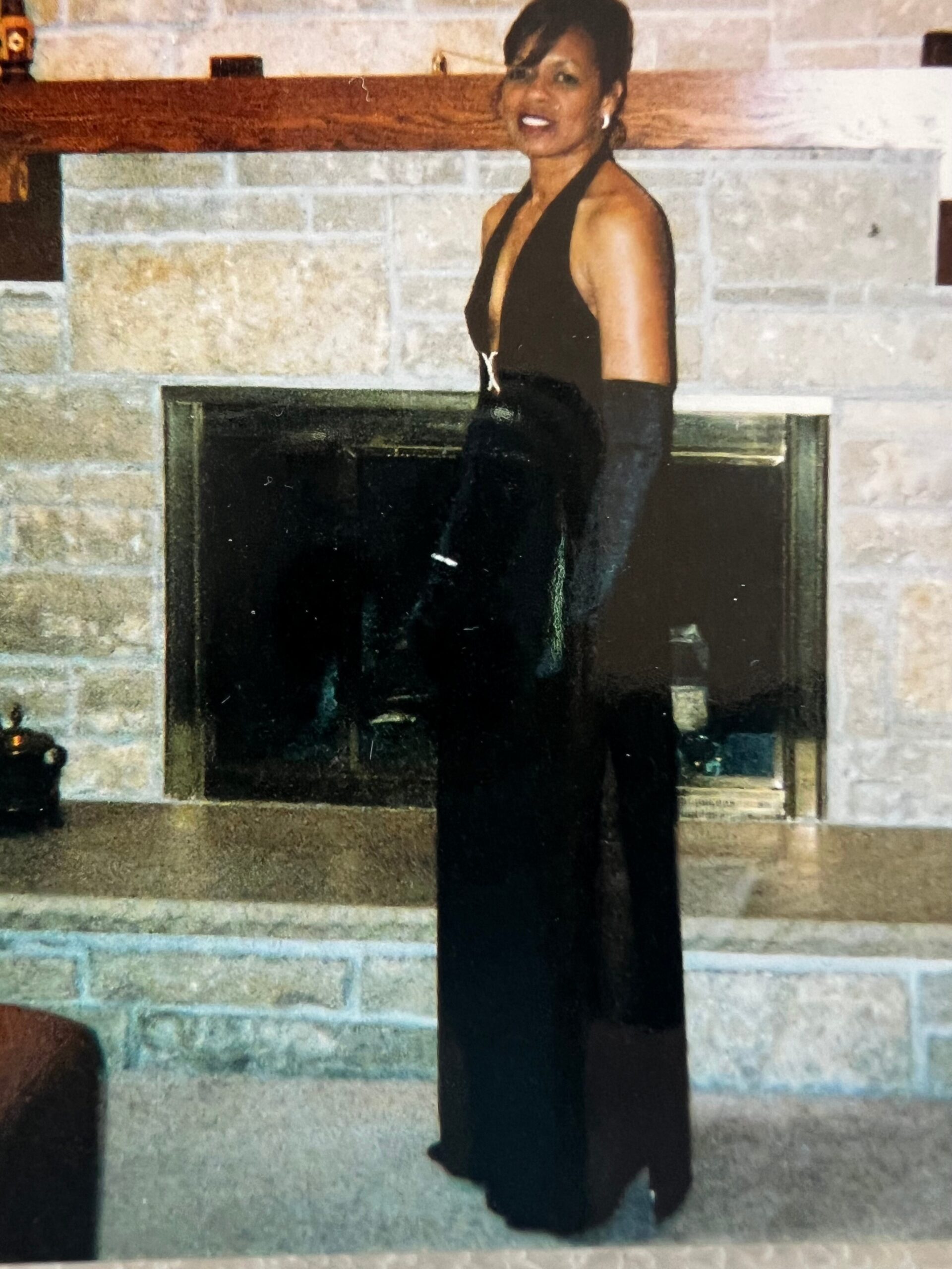 Woman in a black halter gown and long gloves stands by a stone fireplace, smiling at the camera.