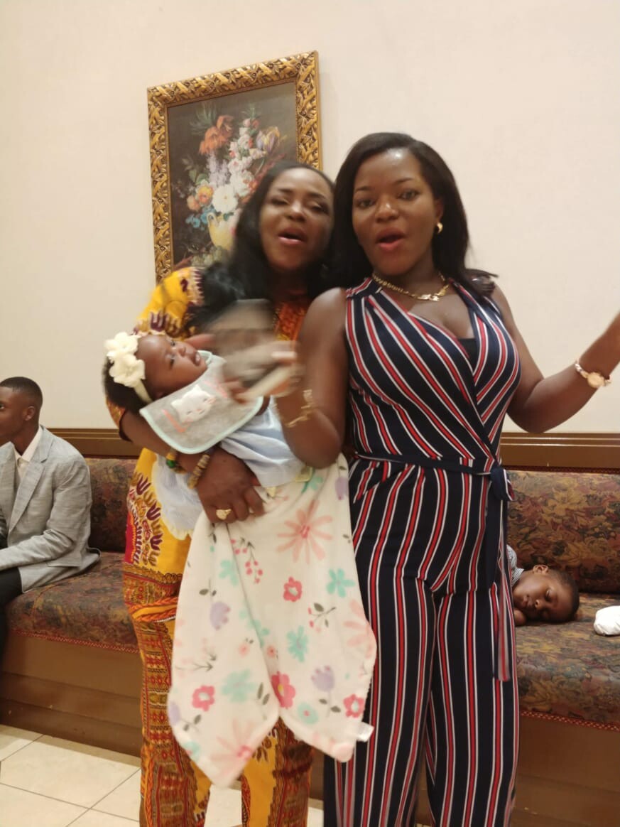 Two women stand indoors with a baby wrapped in a floral blanket, while others sit on a patterned couch in the background.
